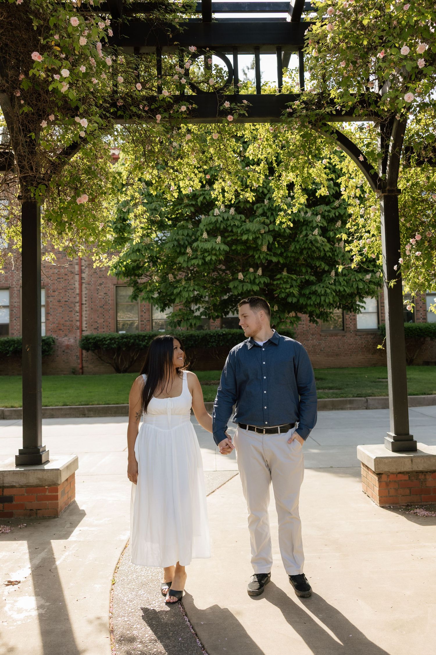 Couple holding hands and walking under a floral pergola, smiling at each other in a sunlit park setting.