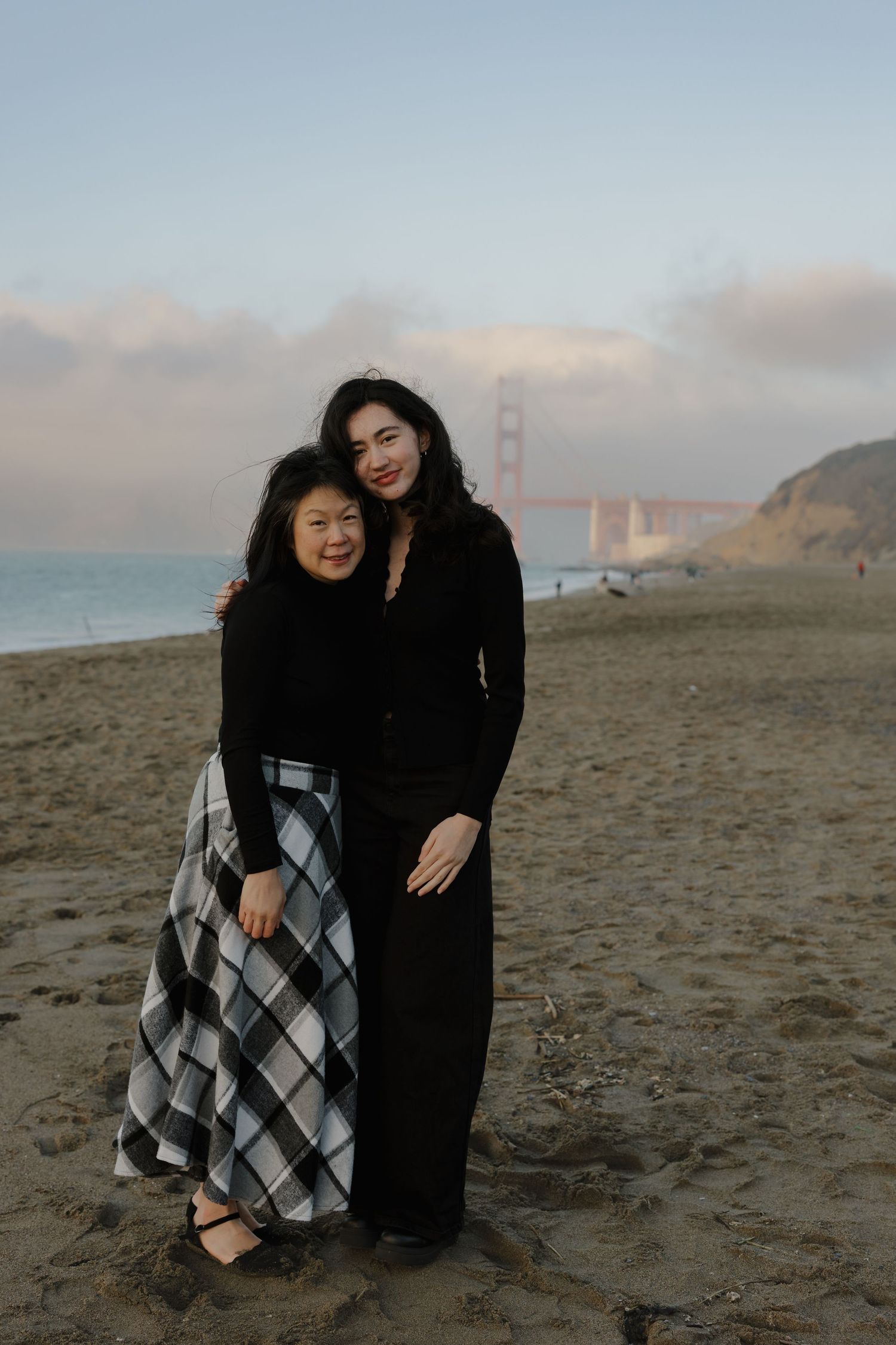 A couple sits together on a sandy beach with the iconic Golden Gate Bridge visible in the background during sunset.