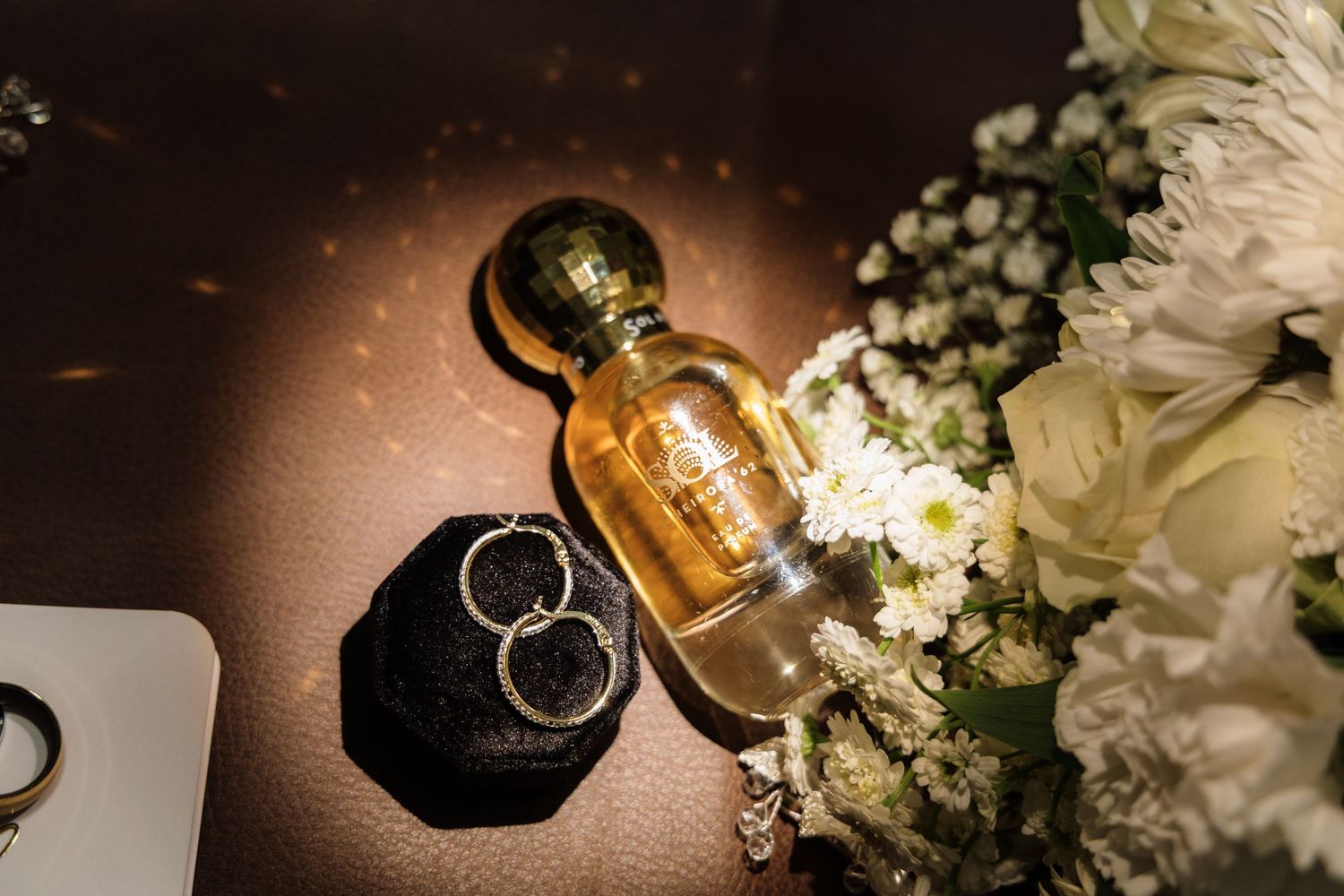 Wedding details including gold perfume bottle, wedding rings, and white flowers arranged on dark surface.