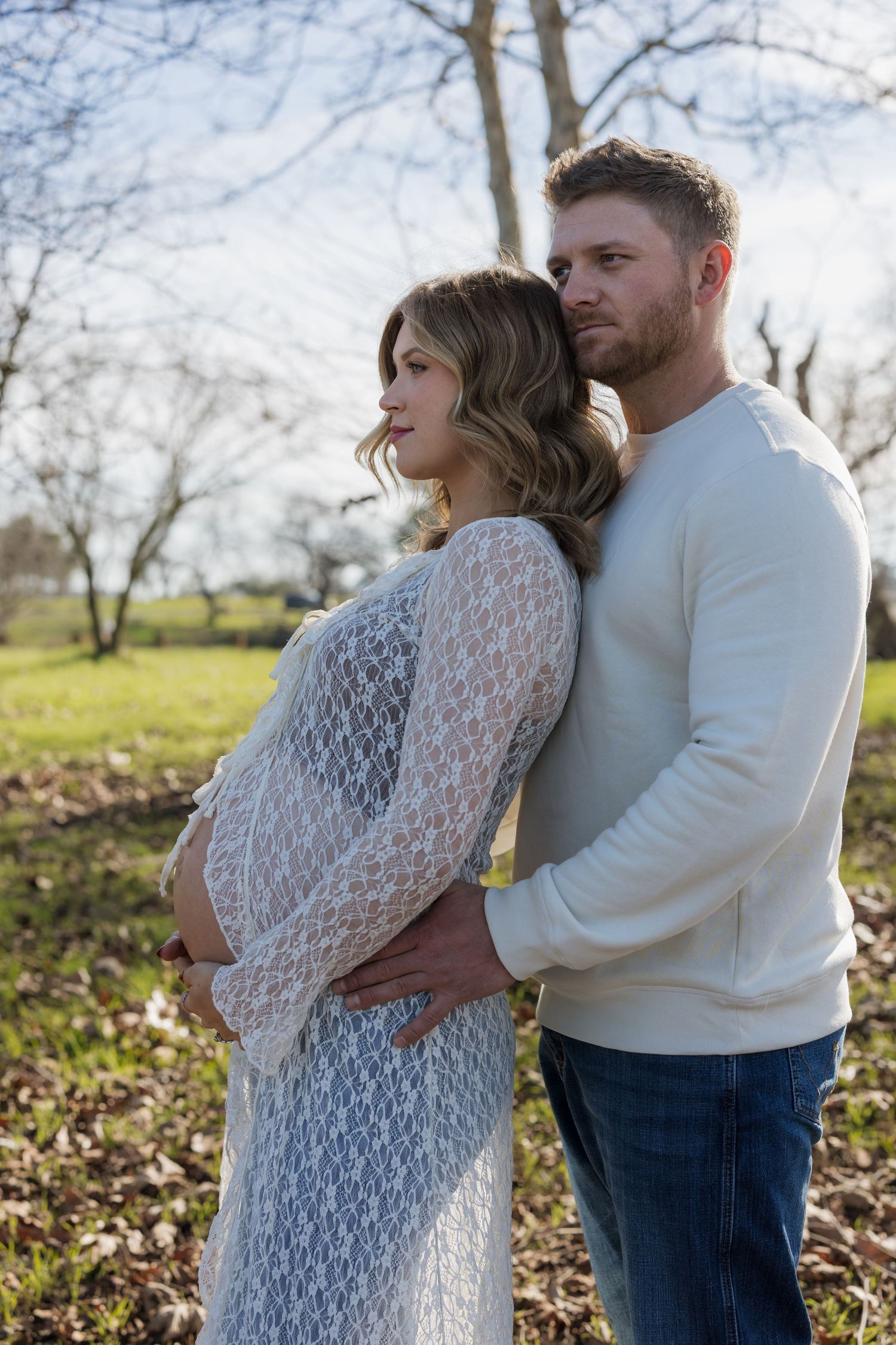 A romantic outdoor maternity photoshoot showing a couple embracing in a sunlit field with bare trees in the background.