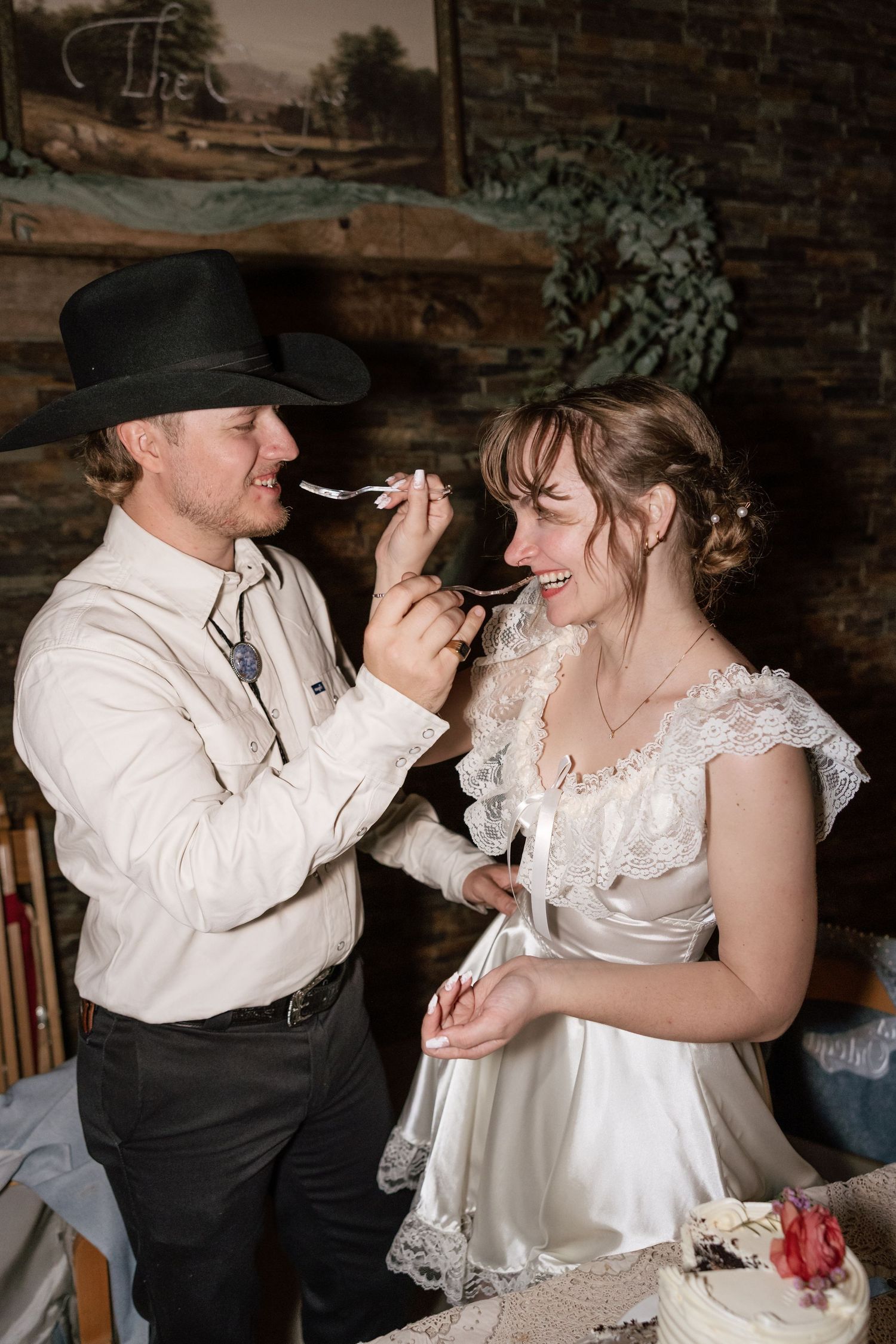 Couple sharing cake during wedding reception in rustic venue with brick wall backdrop.
