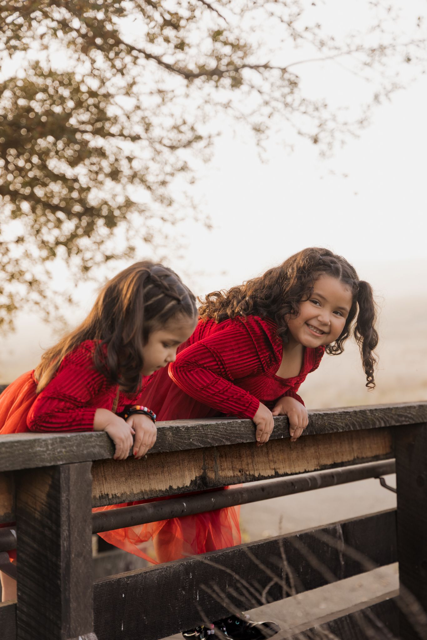 Two children in matching red outfits lean playfully over a wooden railing during a misty autumn day.