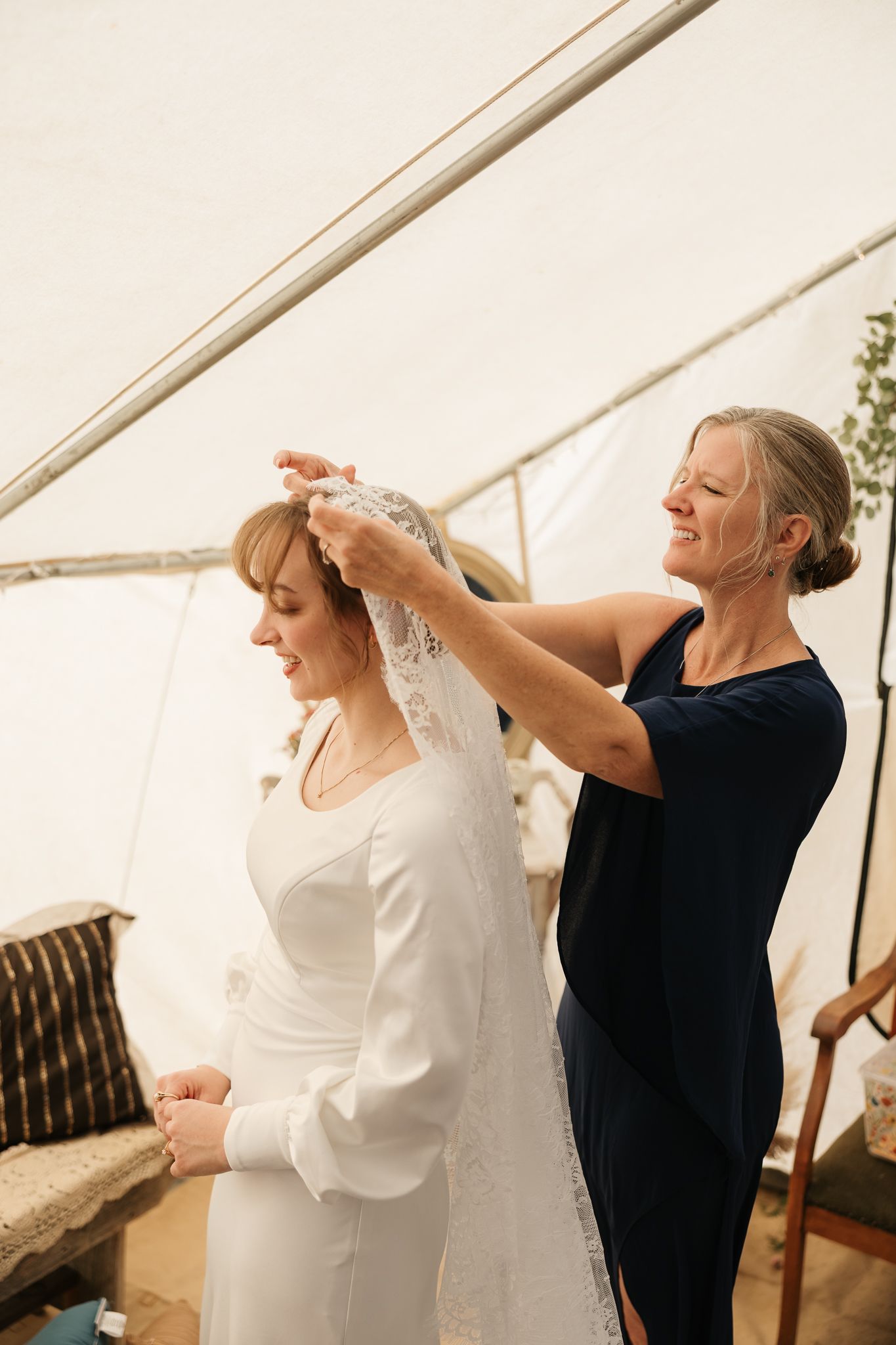 A person in a navy dress helps adjust a long white wedding veil on someone wearing a white long-sleeved bridal gown.