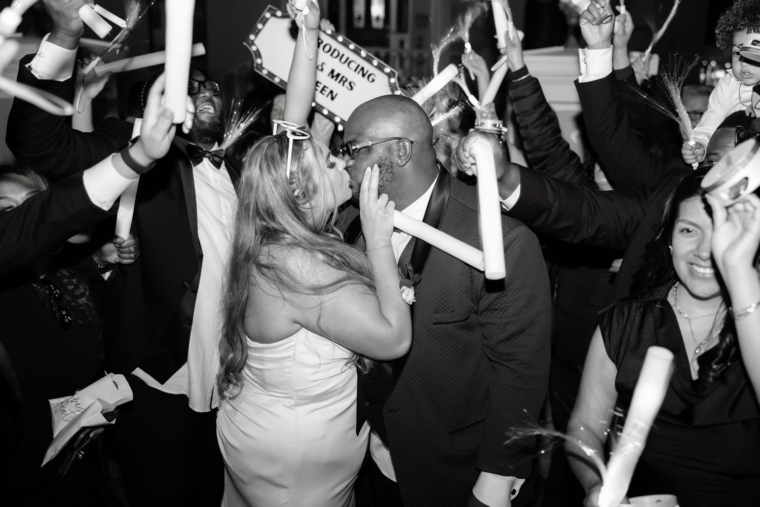 Black and white photo of a bride and groom kissing at their wedding reception surrounded by cheering guests.