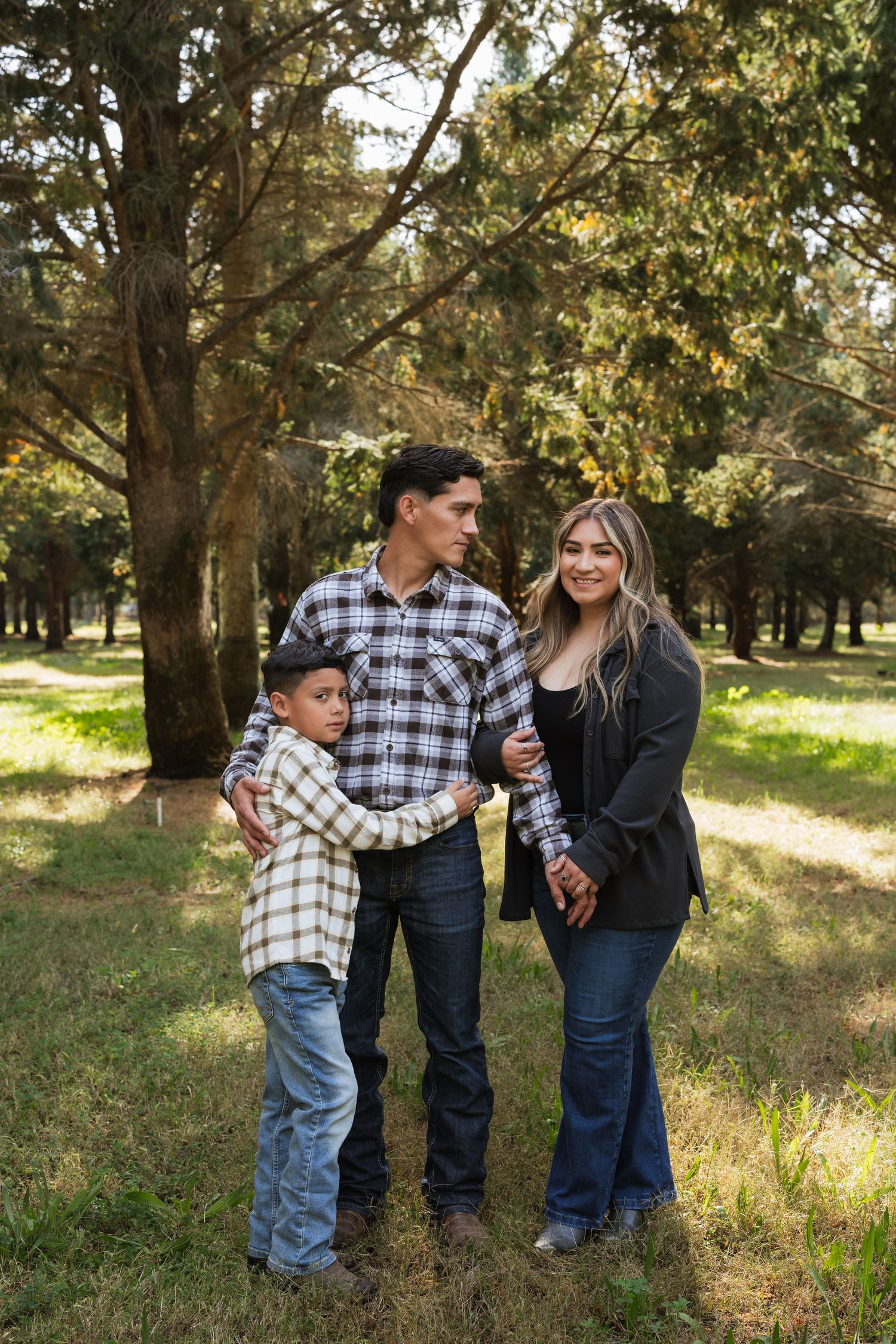 A family shares candid moments together in a sunlit park setting with trees in the background.