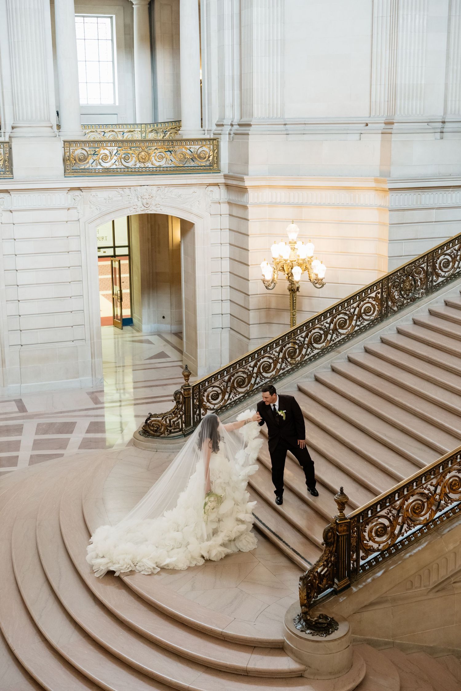 Bride and groom descend grand staircase at San Francisco City Hall, bride's flowing white gown trailing elegantly behind.