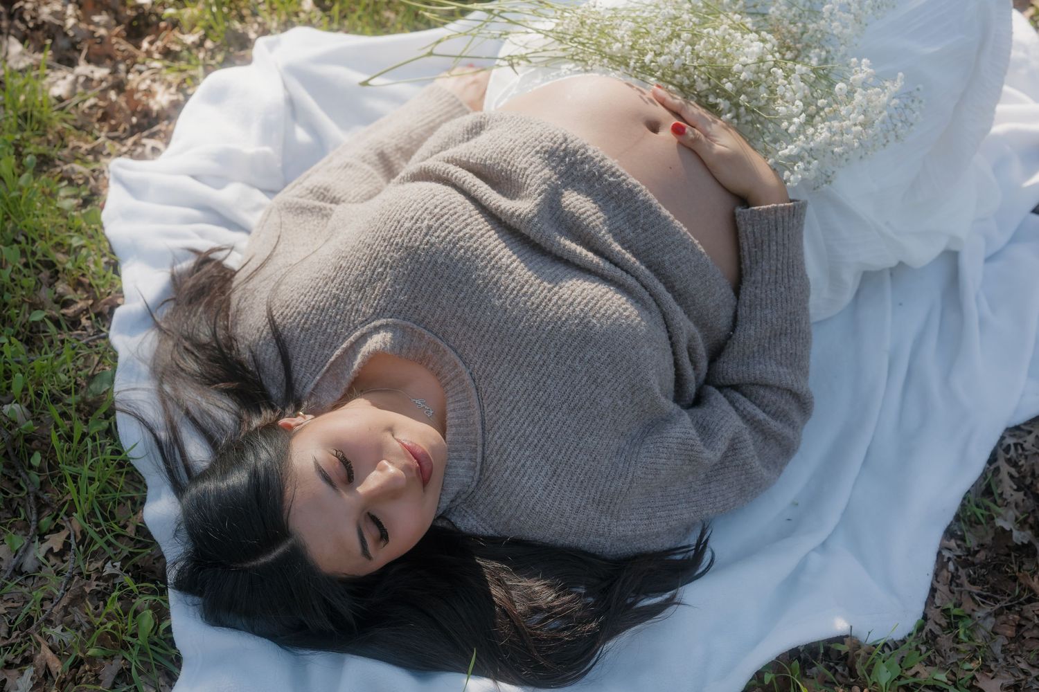 Person lying on white blanket outdoors wearing a cozy gray sweater on a sunny day.