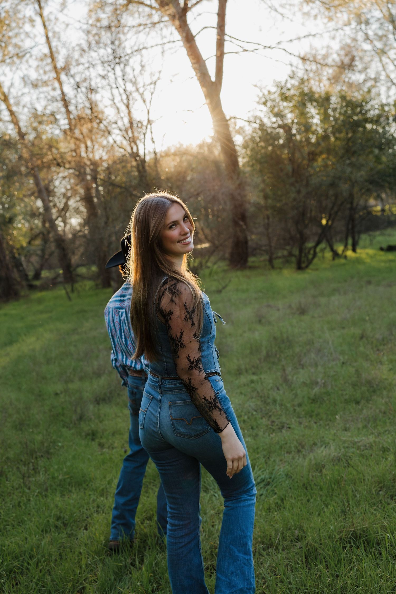 Woman in denim overalls and jean jacket poses playfully in a sunlit grassy field during golden hour.