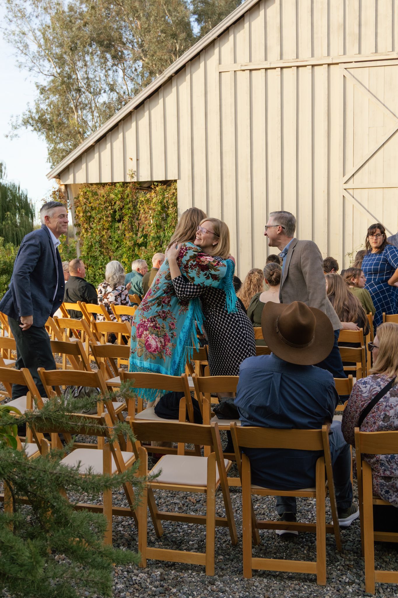 A group of people seated on wooden folding chairs outdoors at a rustic venue.