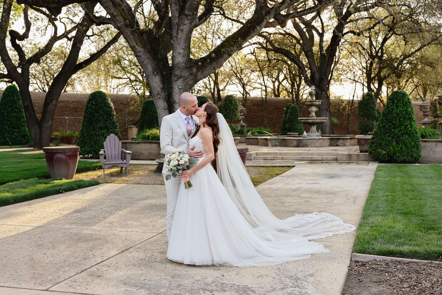 A romantic wedding couple walks together through a garden path lined with trees at golden hour.