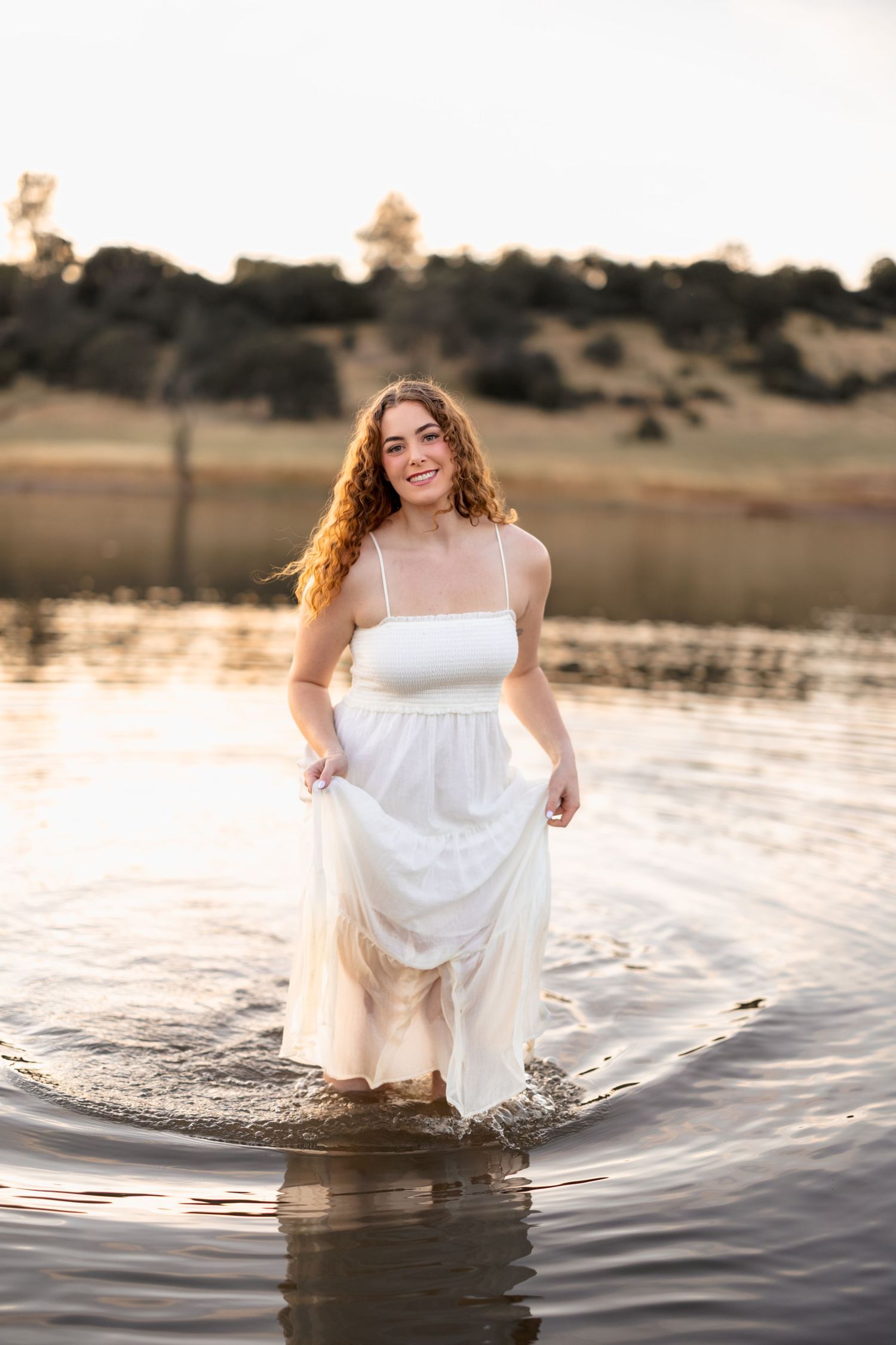 Person in white dress stands in shallow water during sunset.