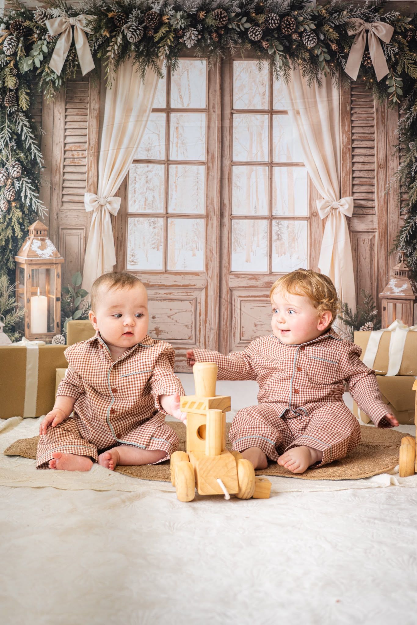 Two babies in matching plaid pajamas sit together near a wooden toy train in a festive Christmas holiday setting.