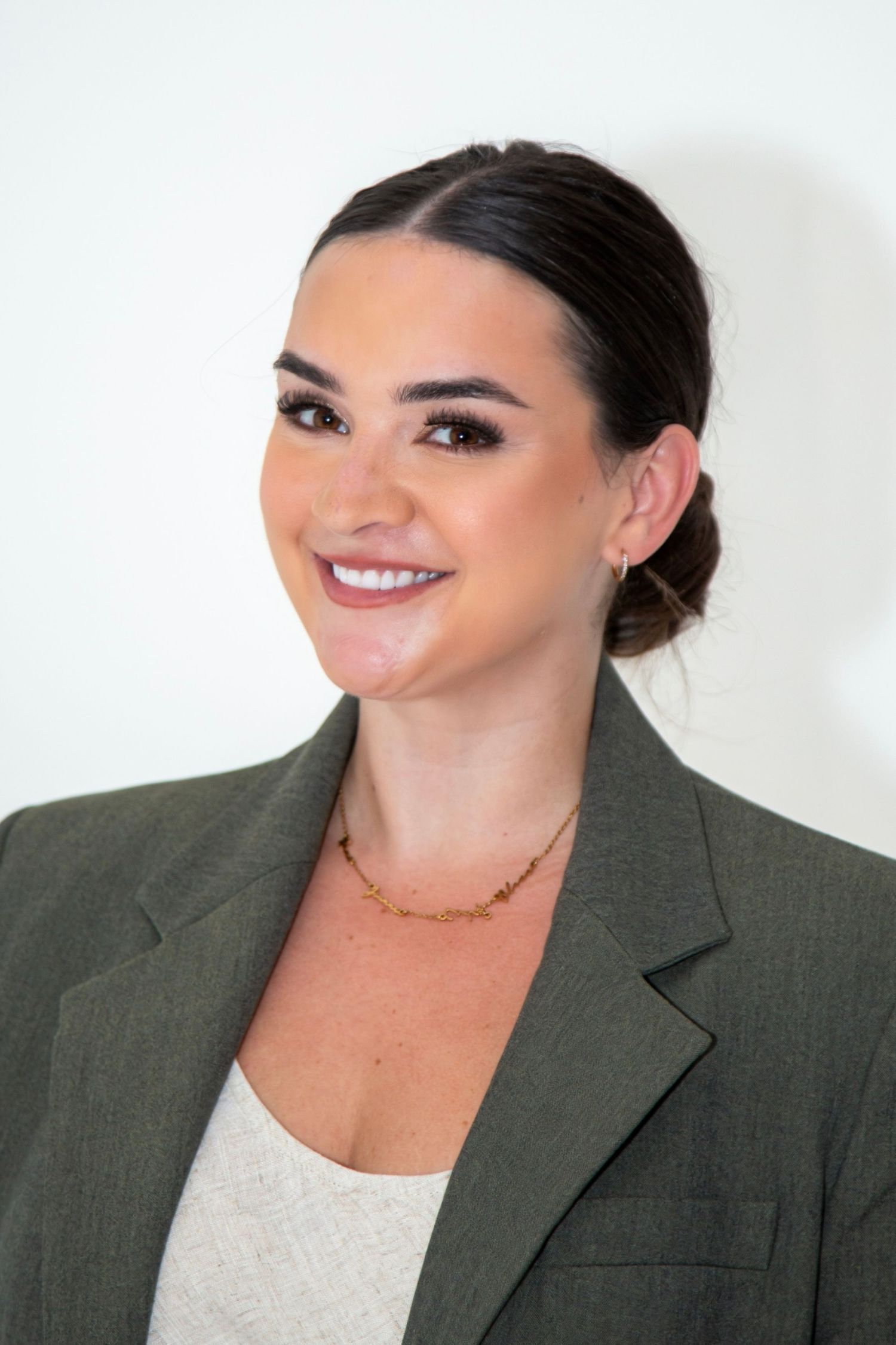 Professional woman in olive blazer with gold necklace, dark hair in updo, smiling confidently against white background.