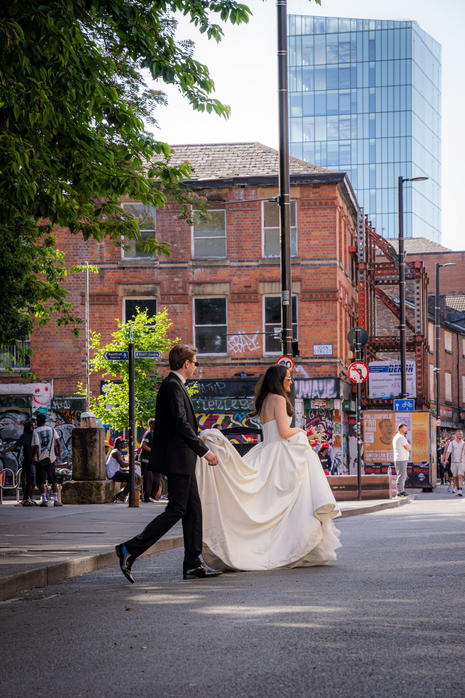 Bride and groom walking hand in hand across a sunny urban street in a vibrant city setting.