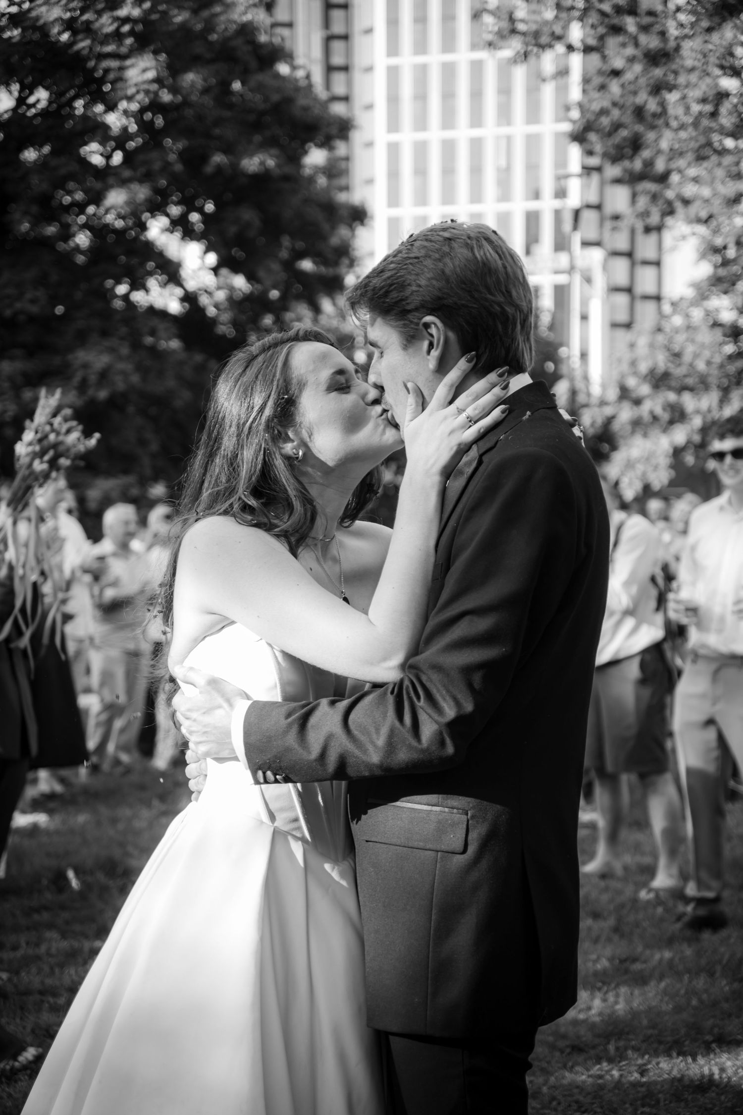 Black and white photo of bride and groom sharing their first kiss outdoors surrounded by wedding guests.