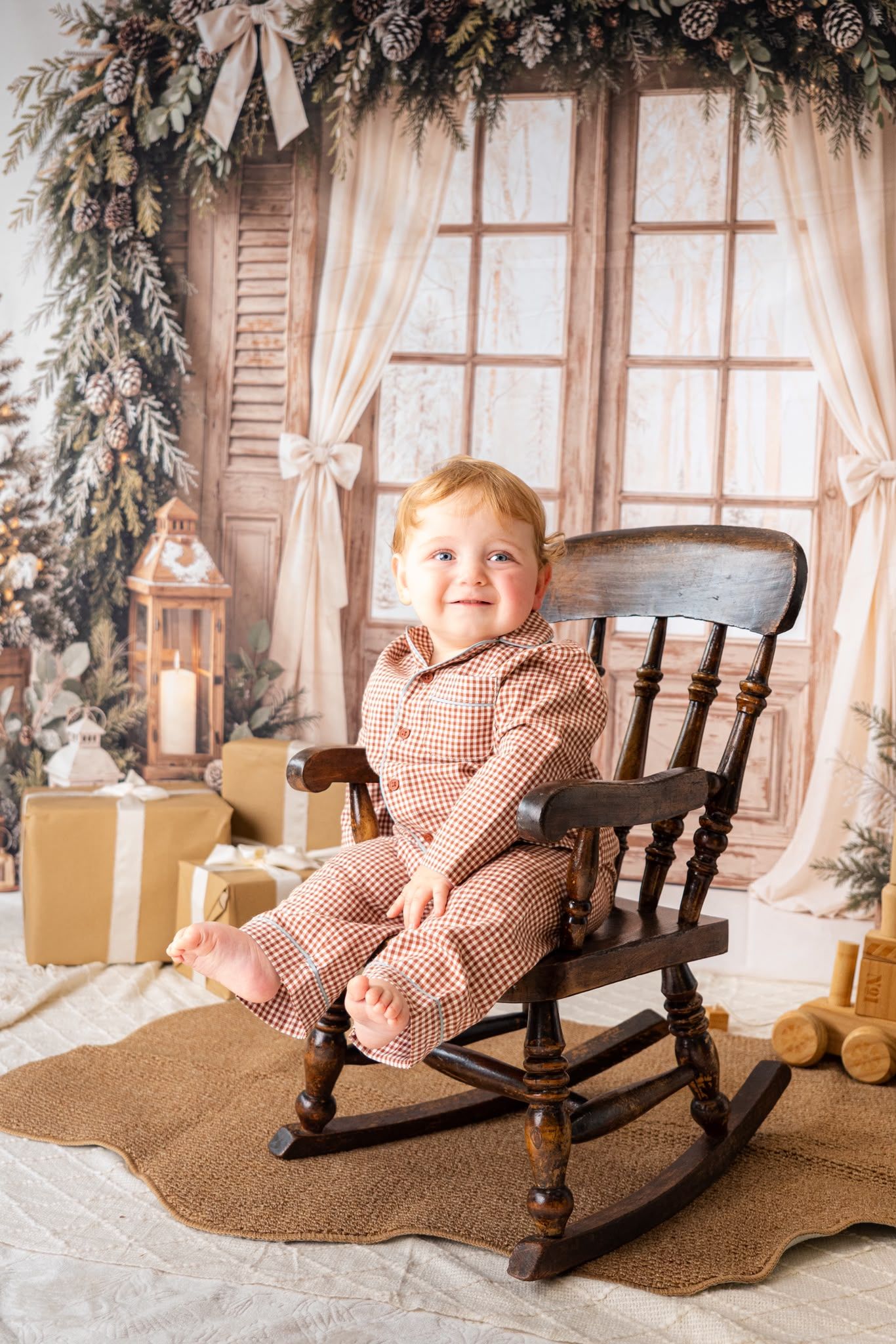 Smiling toddler in red plaid pajamas sitting in a wooden rocking chair with Christmas decorations in the background.