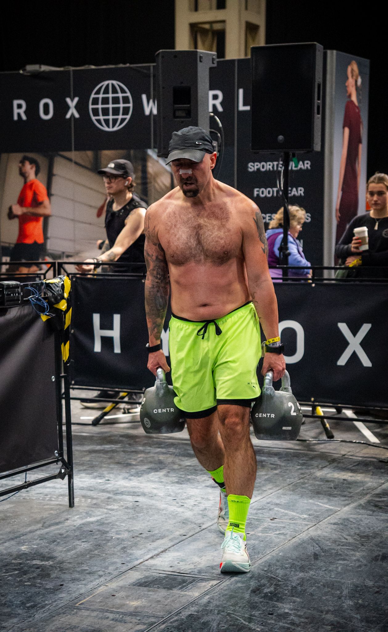 Shirtless male athlete carries kettlebells at a HYROX fitness competition wearing neon green shorts and matching socks.