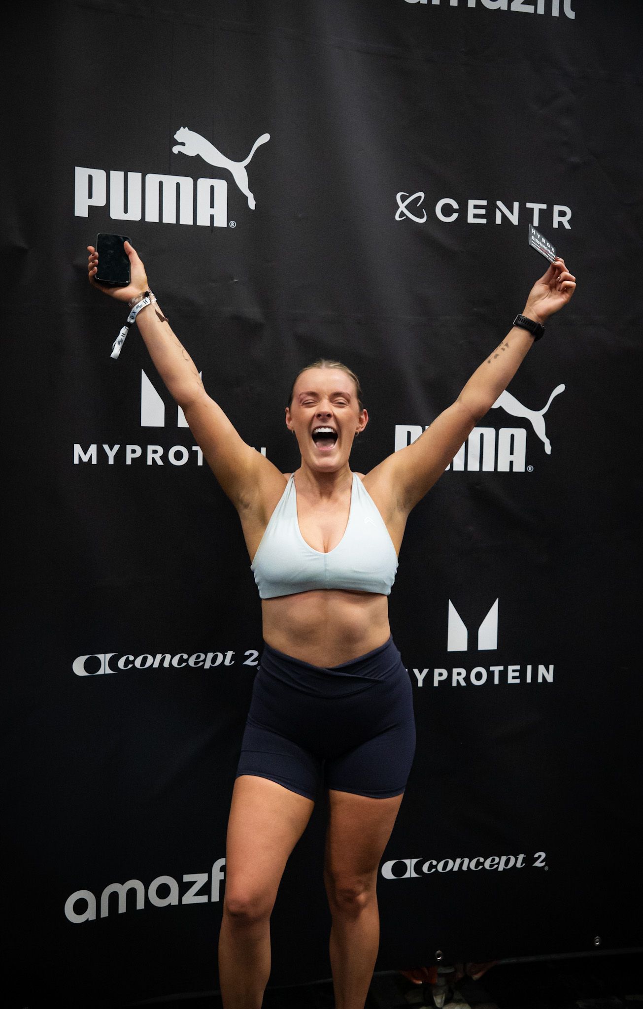 Excited woman in athletic wear celebrates victory at Puma and MyProtein sponsored fitness event, arms raised in triumph.