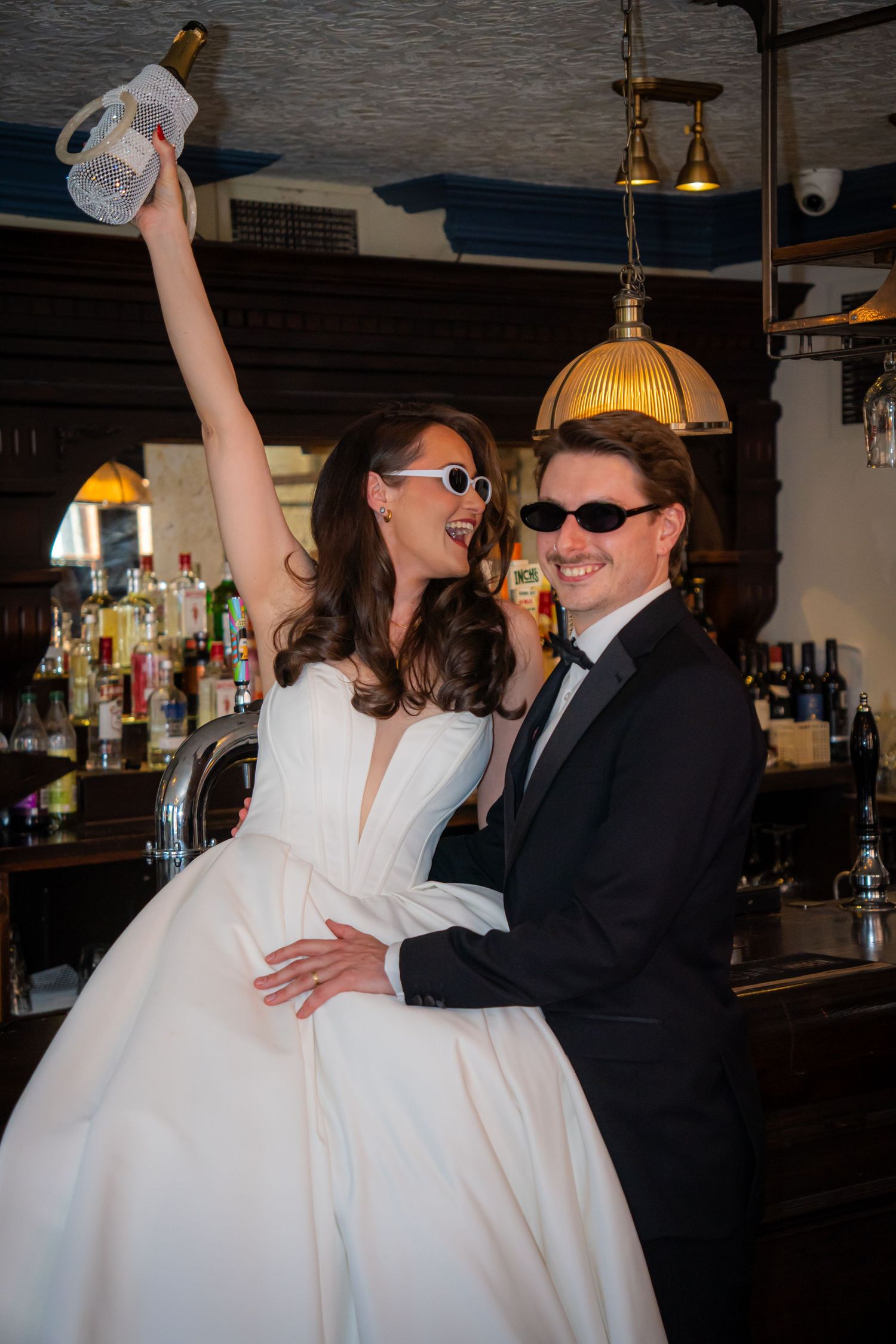 Joyful bride raising champagne bottle at a bar while groom smiles, both wearing sunglasses in playful pose.