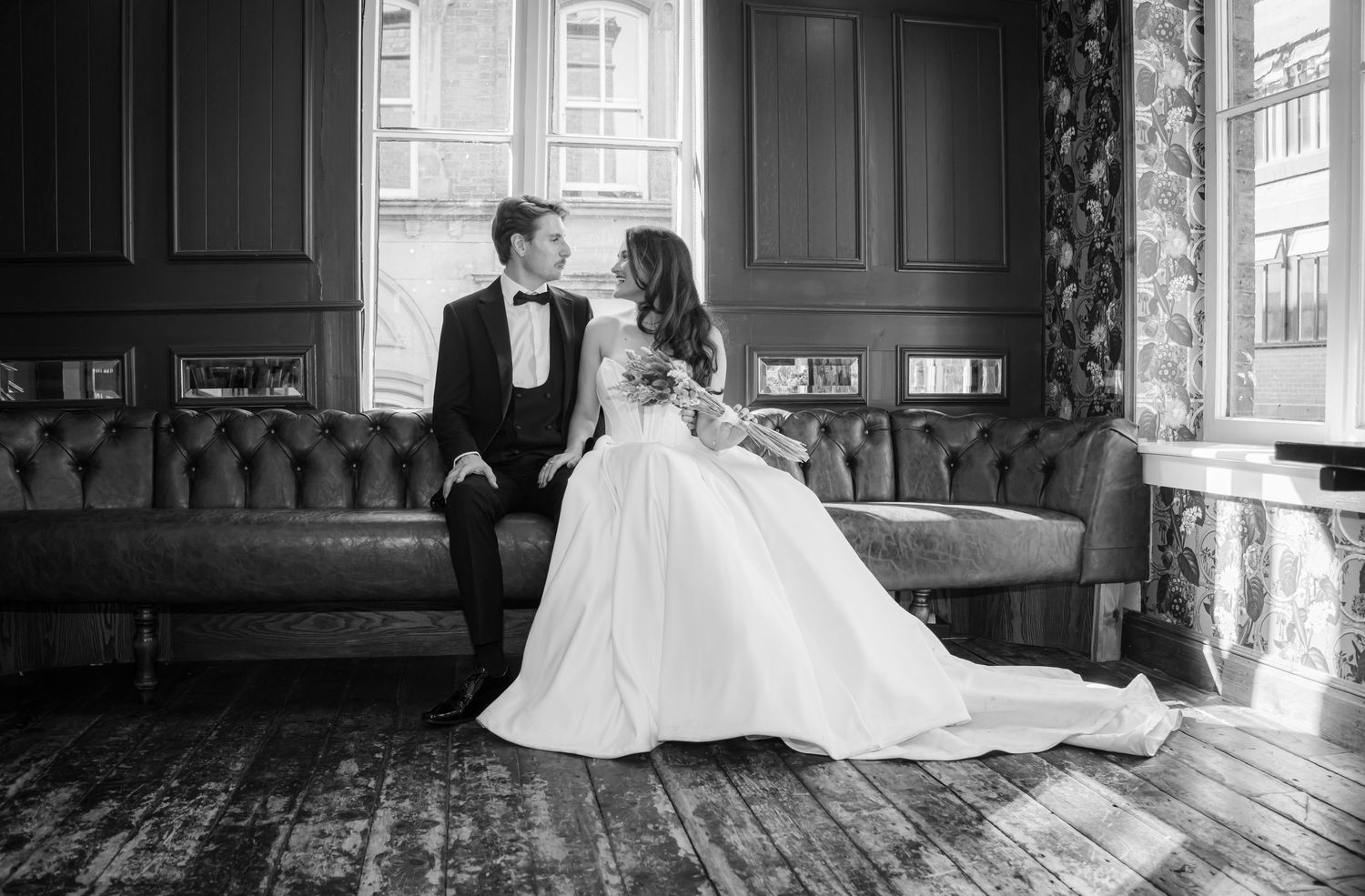 Classic black and white wedding portrait of couple sitting closely together on a tufted leather couch.