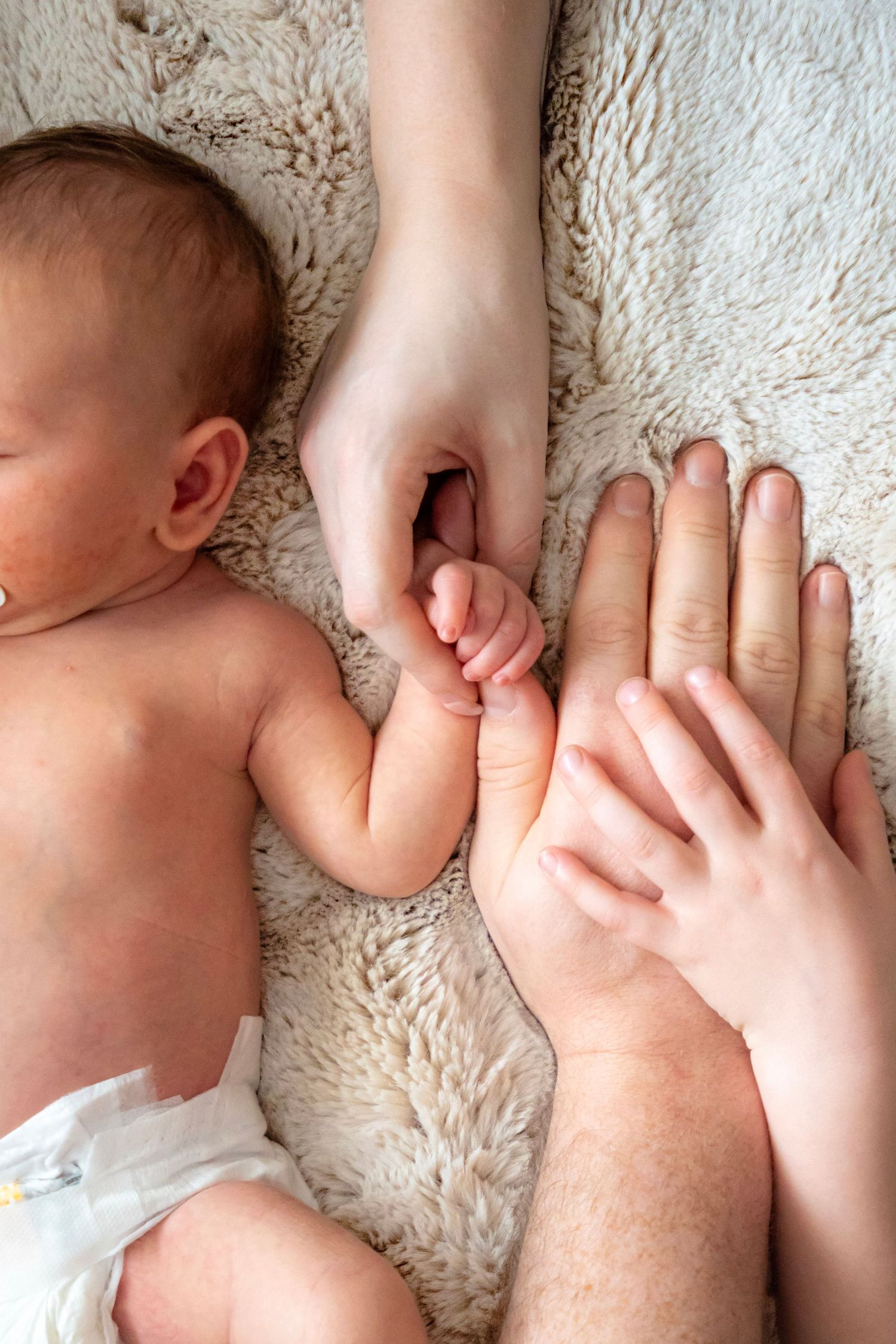 Newborn baby lying on fluffy blanket while two adult hands gently hold the infant's tiny hand.