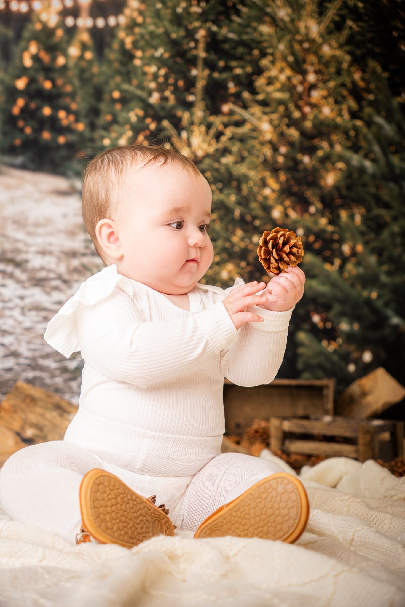 Cute baby in white outfit holding a pine cone, sitting on blanket near Christmas tree with warm bokeh lights.