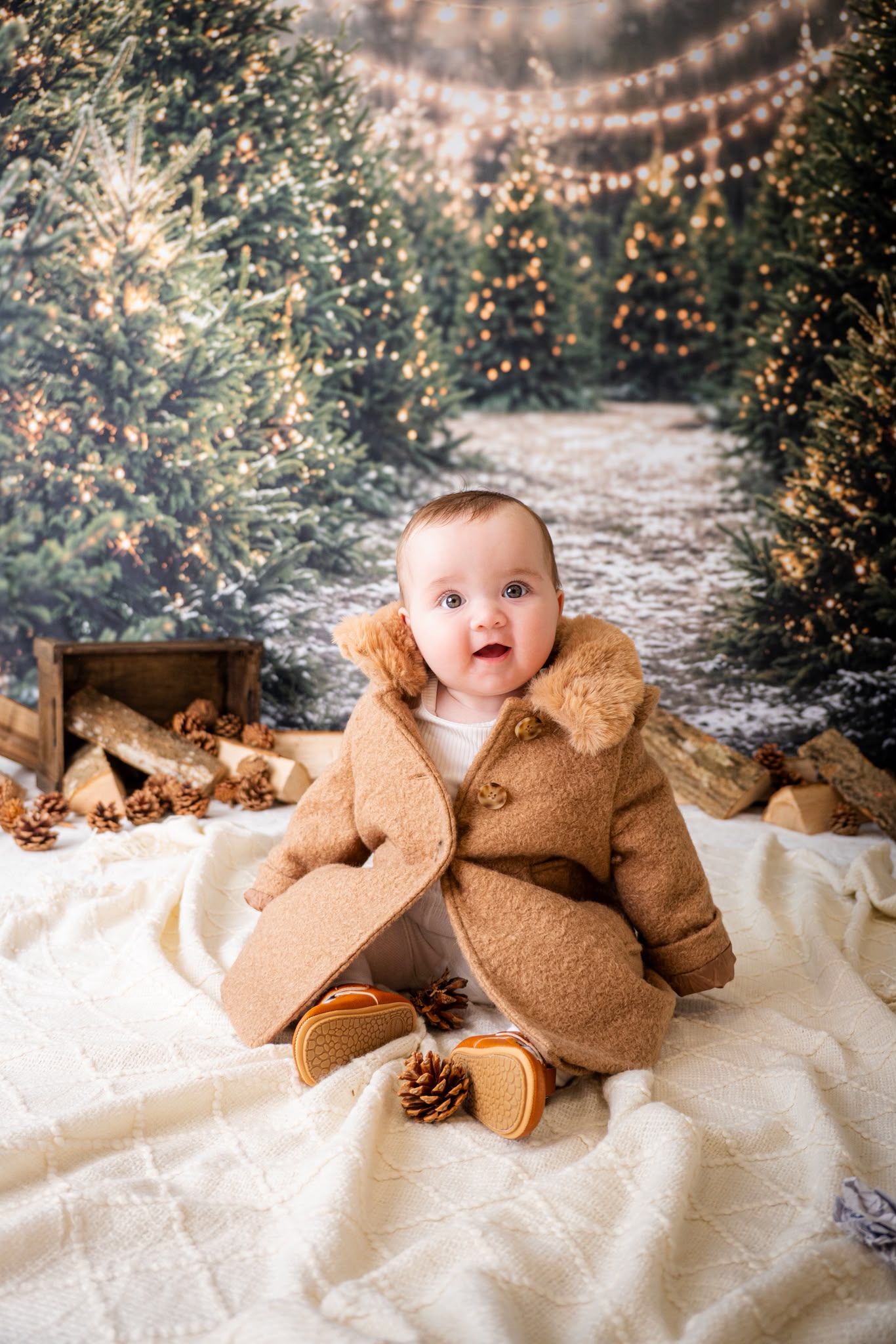 Adorable baby in brown bear snowsuit sits on white blanket surrounded by pine cones and Christmas trees with warm lights.