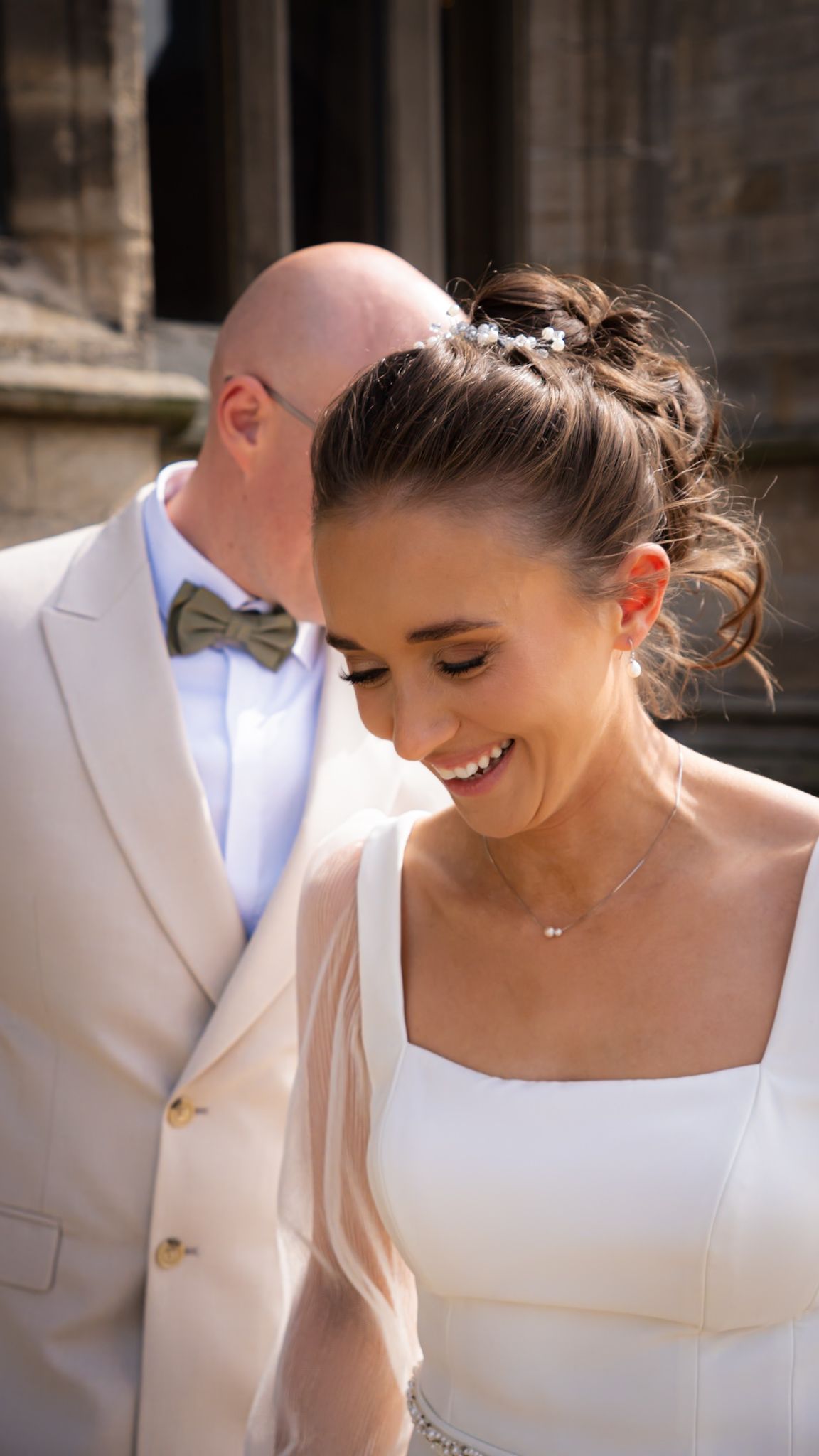 Smiling bride with updo hairstyle and hair accessory, groom in beige suit with bow tie kissing her head.