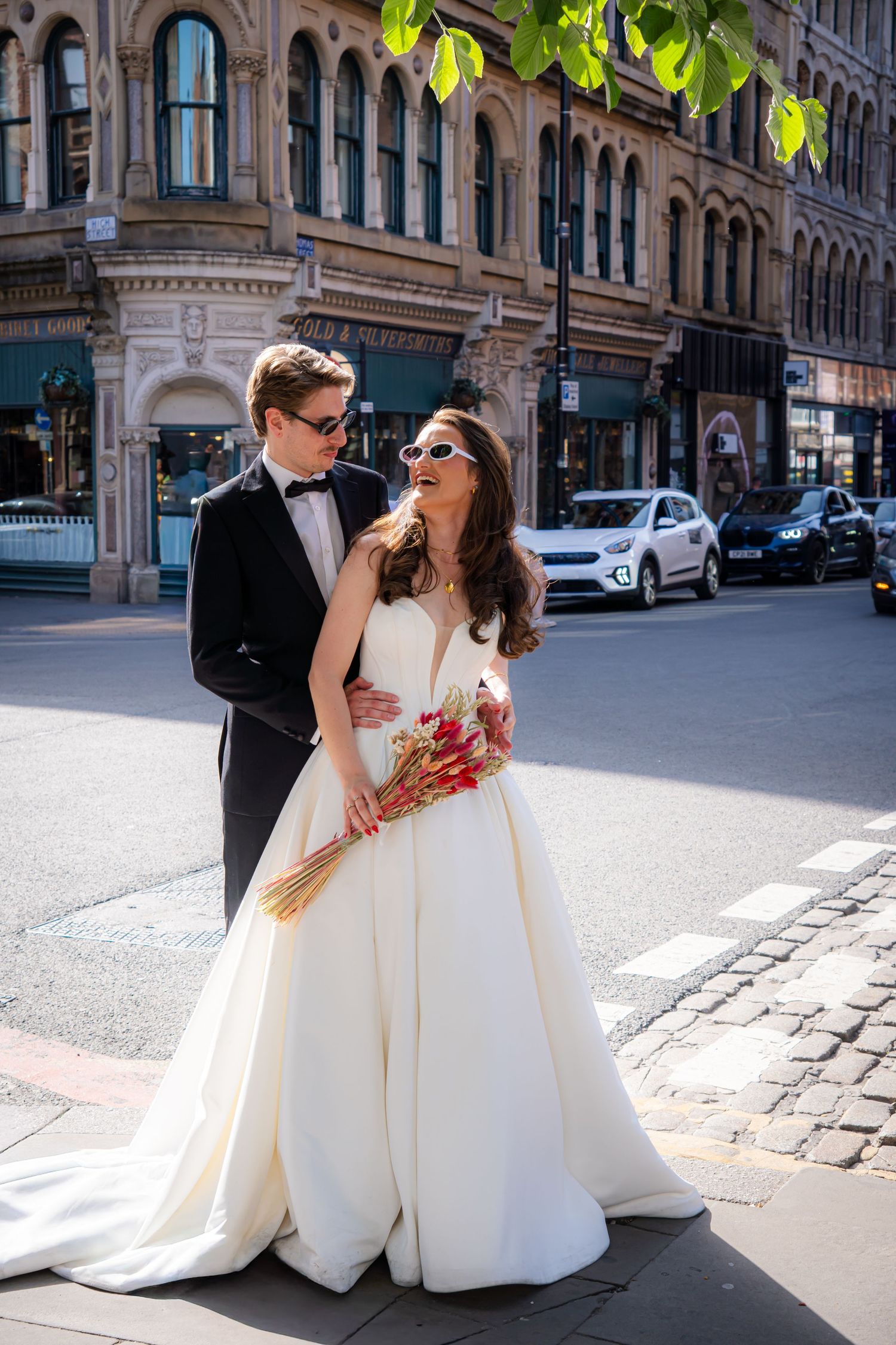 Bride holding a red bouquet and groom embracing on a city street corner on their wedding day.
