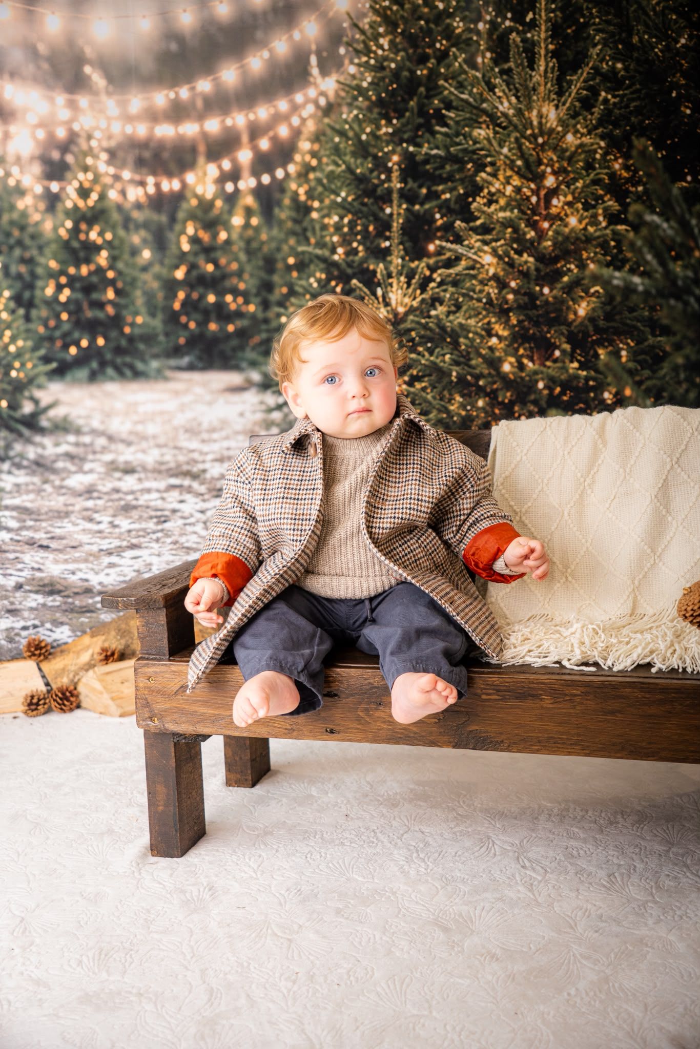 Toddler boy in plaid coat sits on wooden bench surrounded by Christmas trees and string lights in snowy holiday setting.