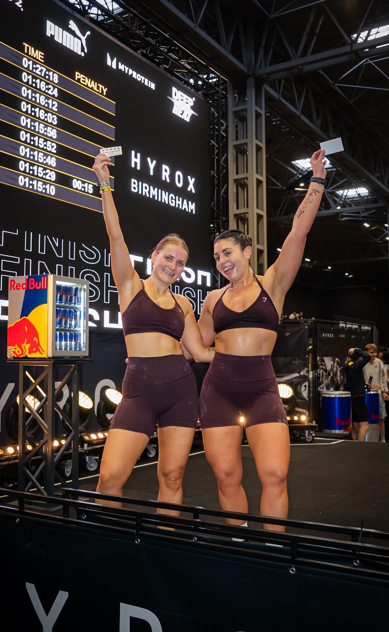 Two women in matching burgundy athletic wear celebrate their finish at HYROX Birmingham fitness race event.