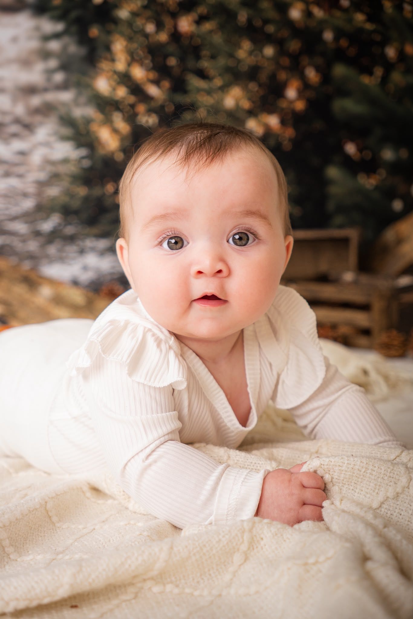 Adorable baby in white outfit lying on a cream blanket, looking up with wide eyes near a Christmas tree.