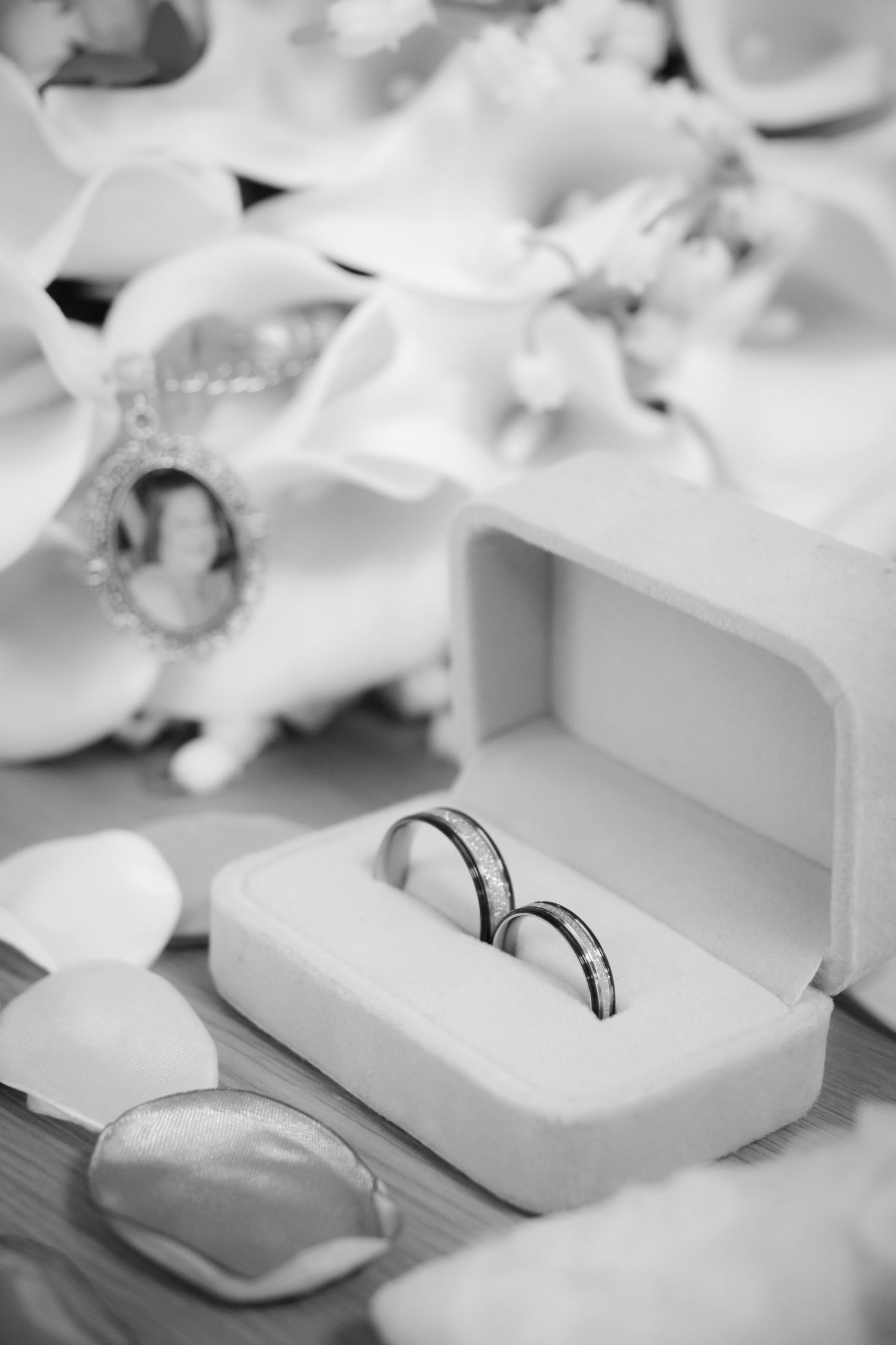 Black and white photo of two gold wedding bands in white ring box with flower petals and decorations.
