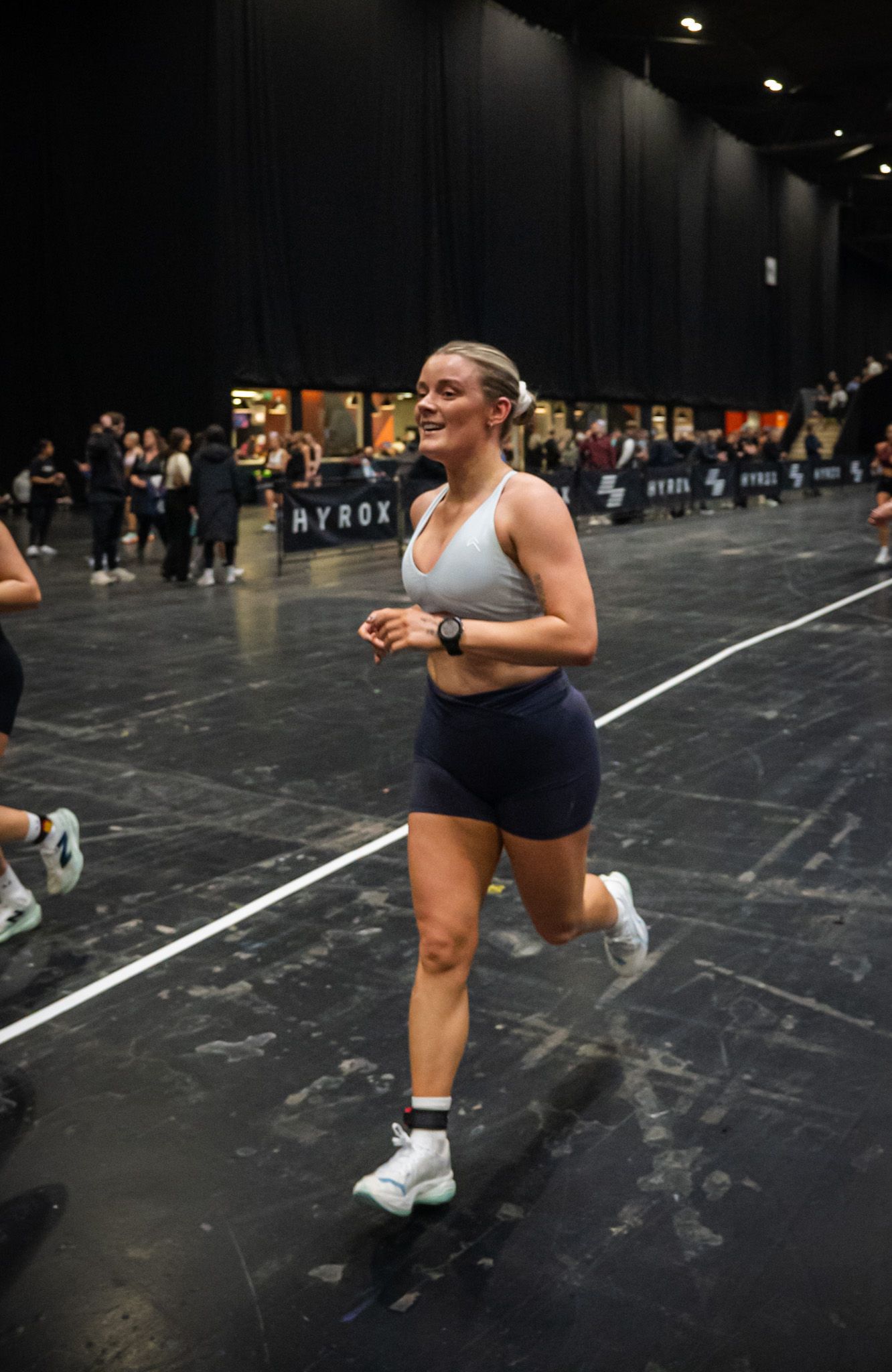 Woman running and smiling at a HYROX fitness competition event, wearing a gray sports bra and navy shorts.