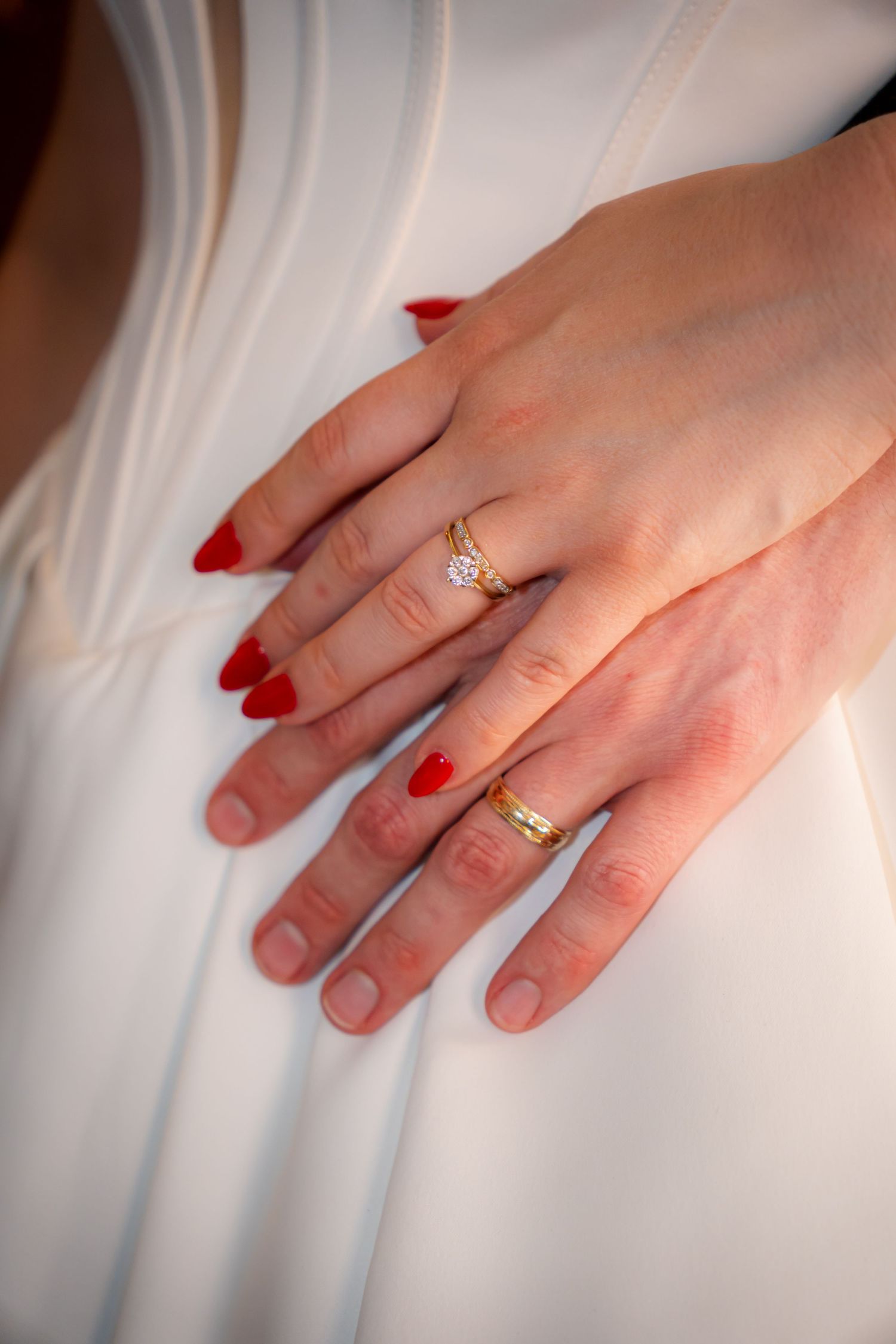 Close-up of bride and groom hands showing gold wedding and engagement rings, with red manicured nails on white dress.