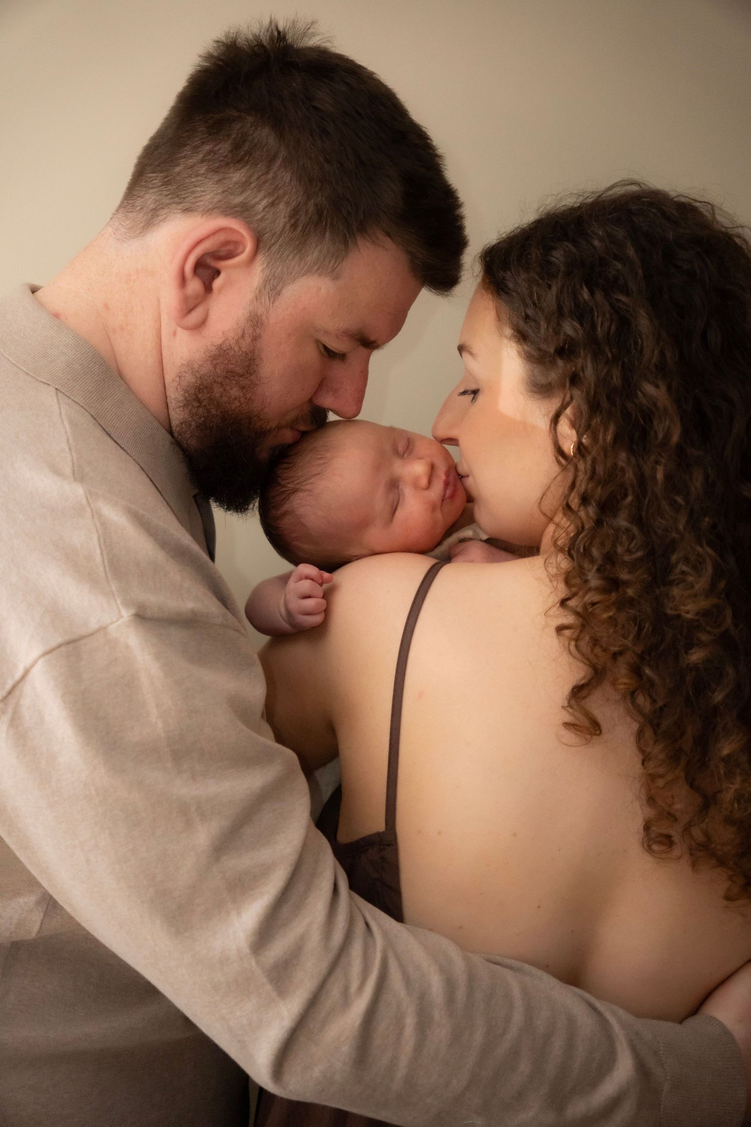 Parents tenderly kissing their sleeping newborn baby together in a warm, intimate family portrait.