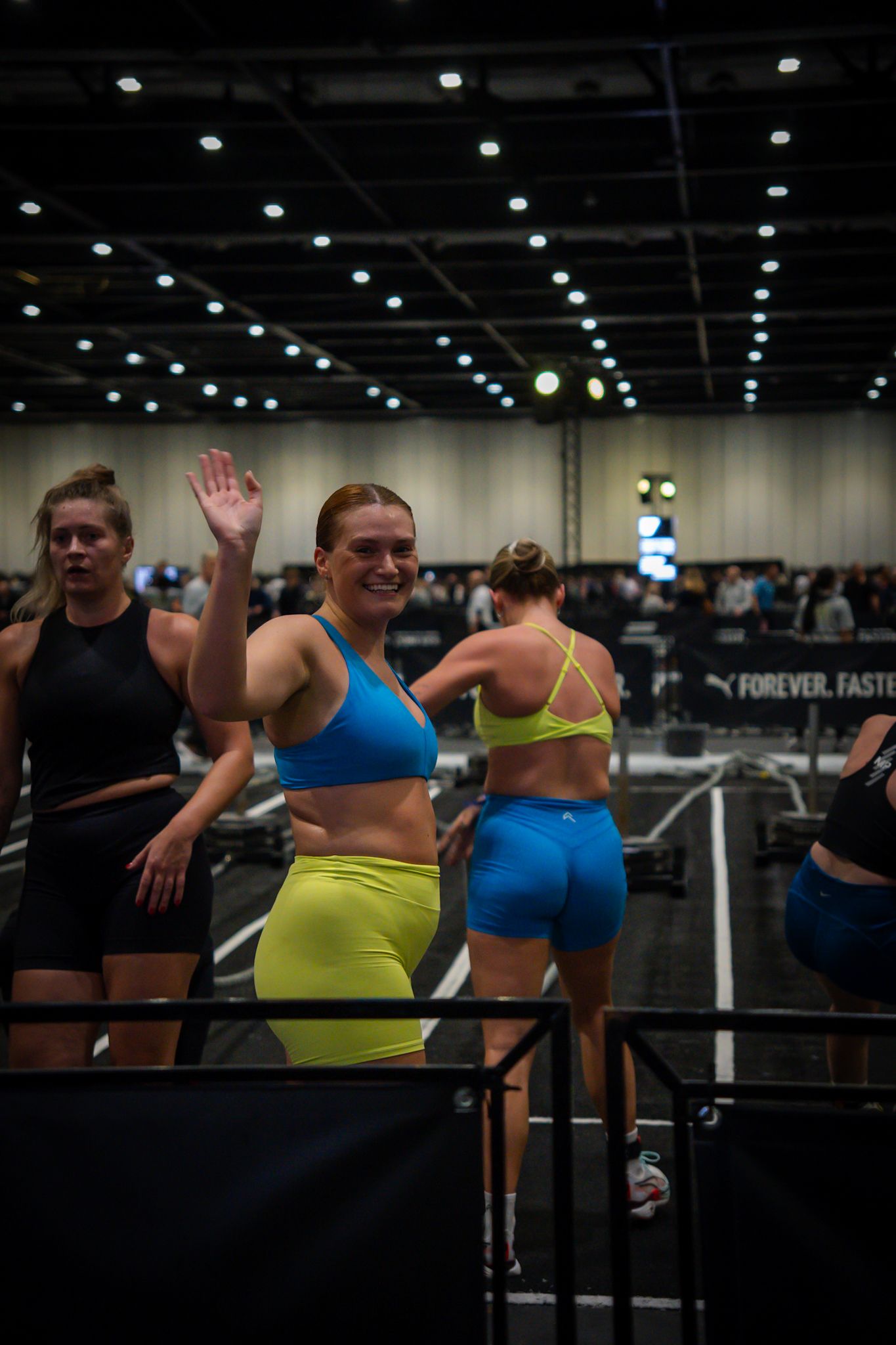 Smiling female athlete in blue sports bra and yellow shorts waving at a fitness competition event indoors.
