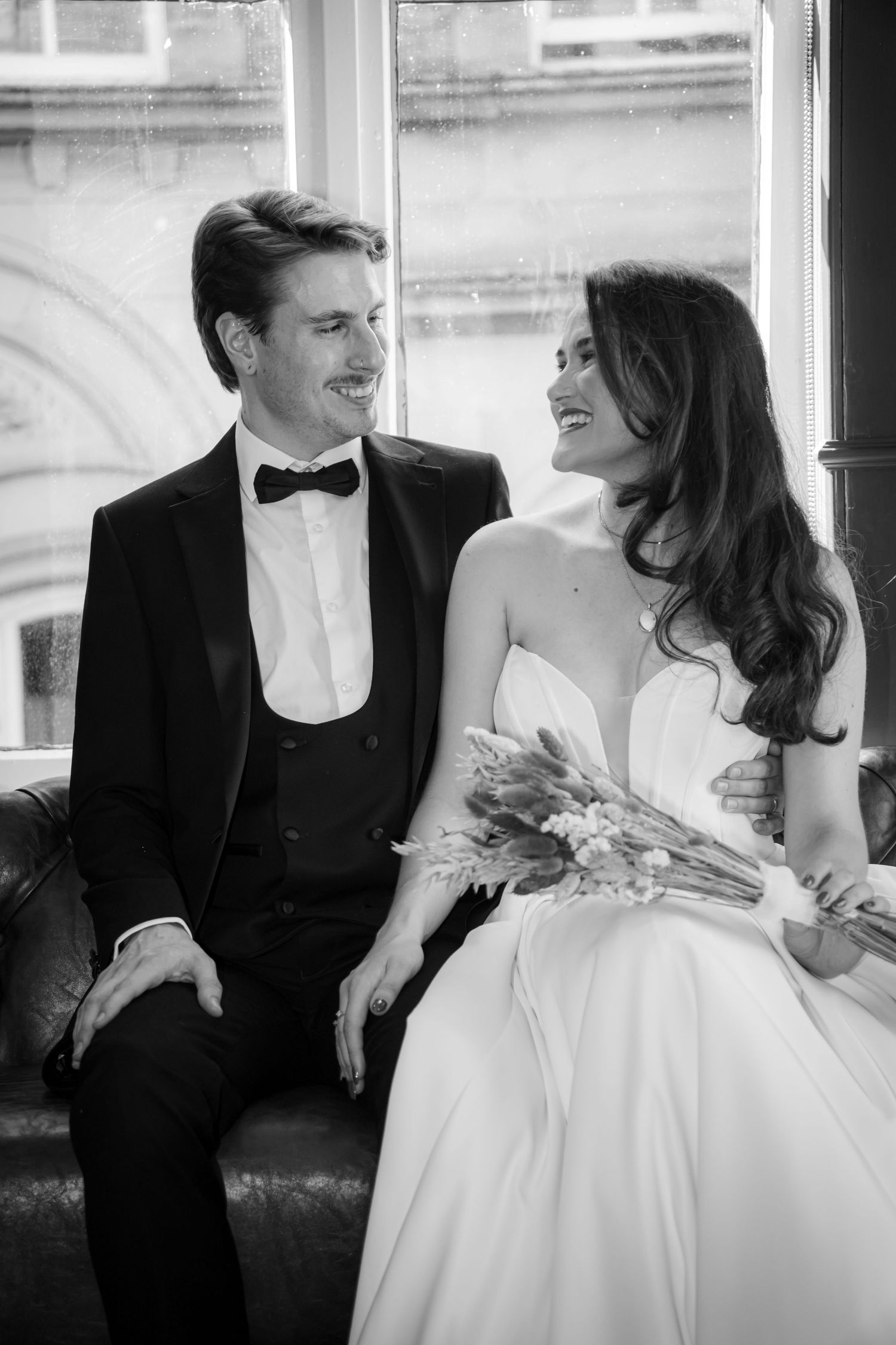 Black and white photo of laughing bride and groom sitting together by a window holding a dried bouquet.