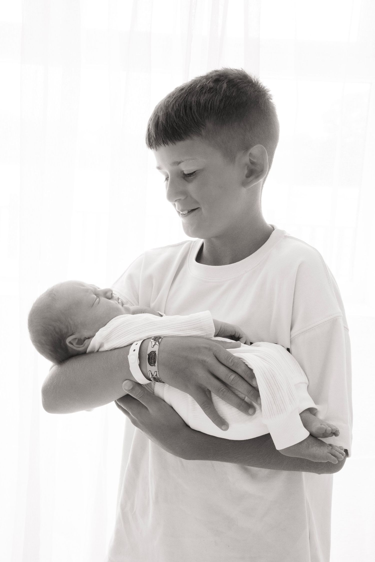 Black and white photo of a smiling boy gently cradling a sleeping newborn baby in his arms.