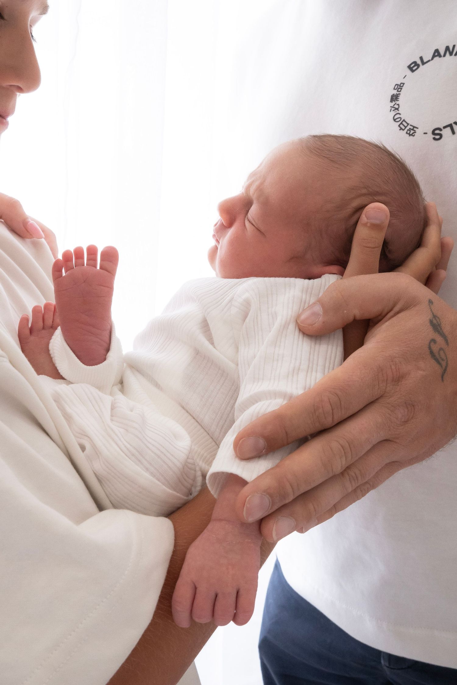 Newborn baby sleeping peacefully while being held by parents in white clothing, tiny feet and hands visible.