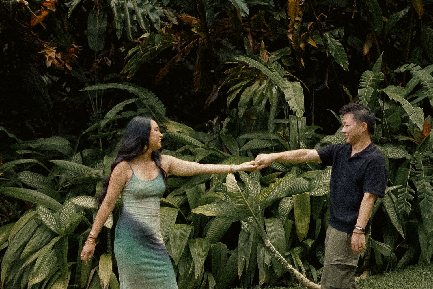 Couple holding hands in a lush tropical garden, woman in green dress and man in black shirt smiling outdoors.