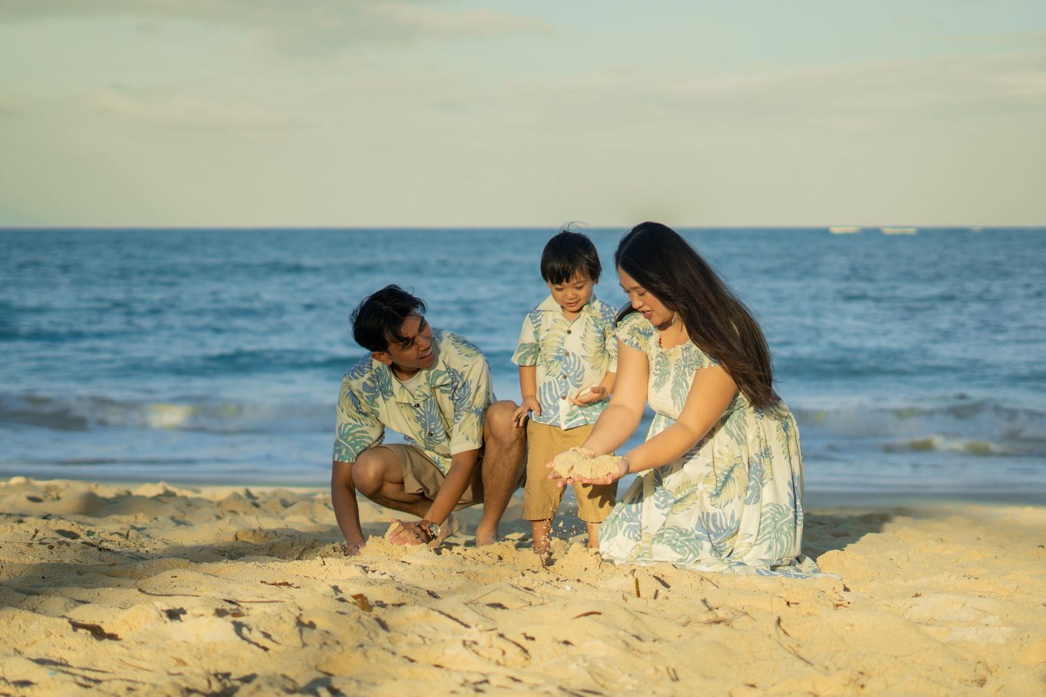 Young family with toddler playing in sand together on sunny Hawaiian beach with ocean backdrop.
