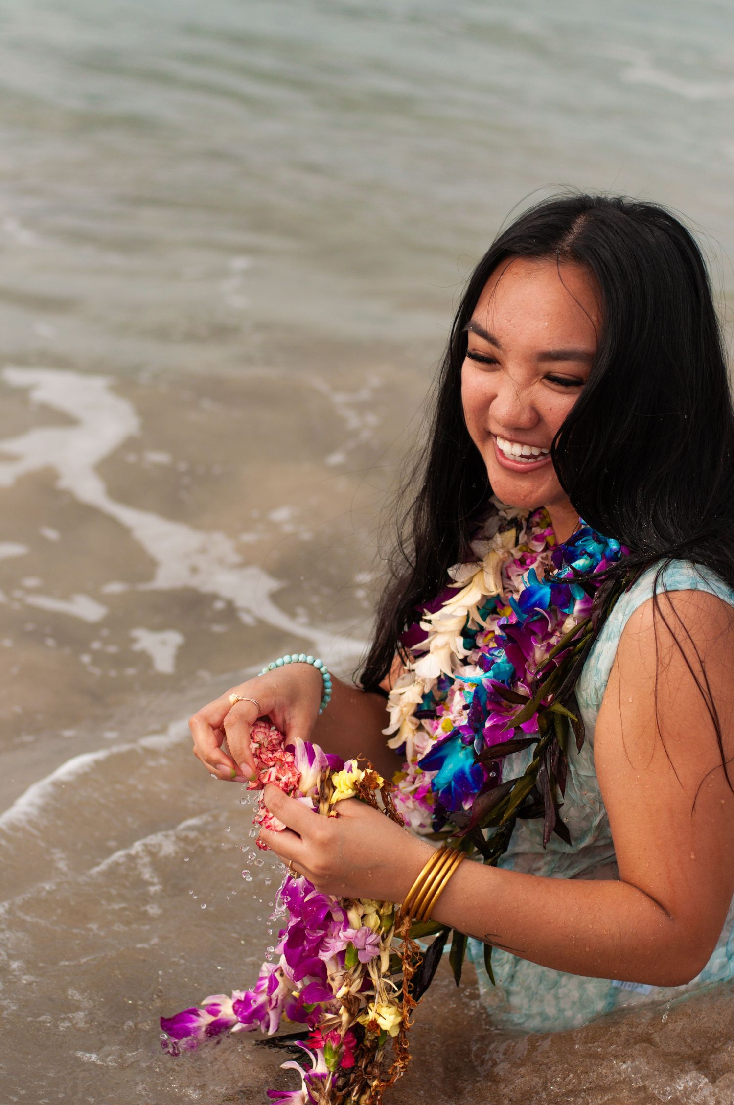 Smiling young woman wearing colorful Hawaiian lei standing in ocean surf on tropical beach.