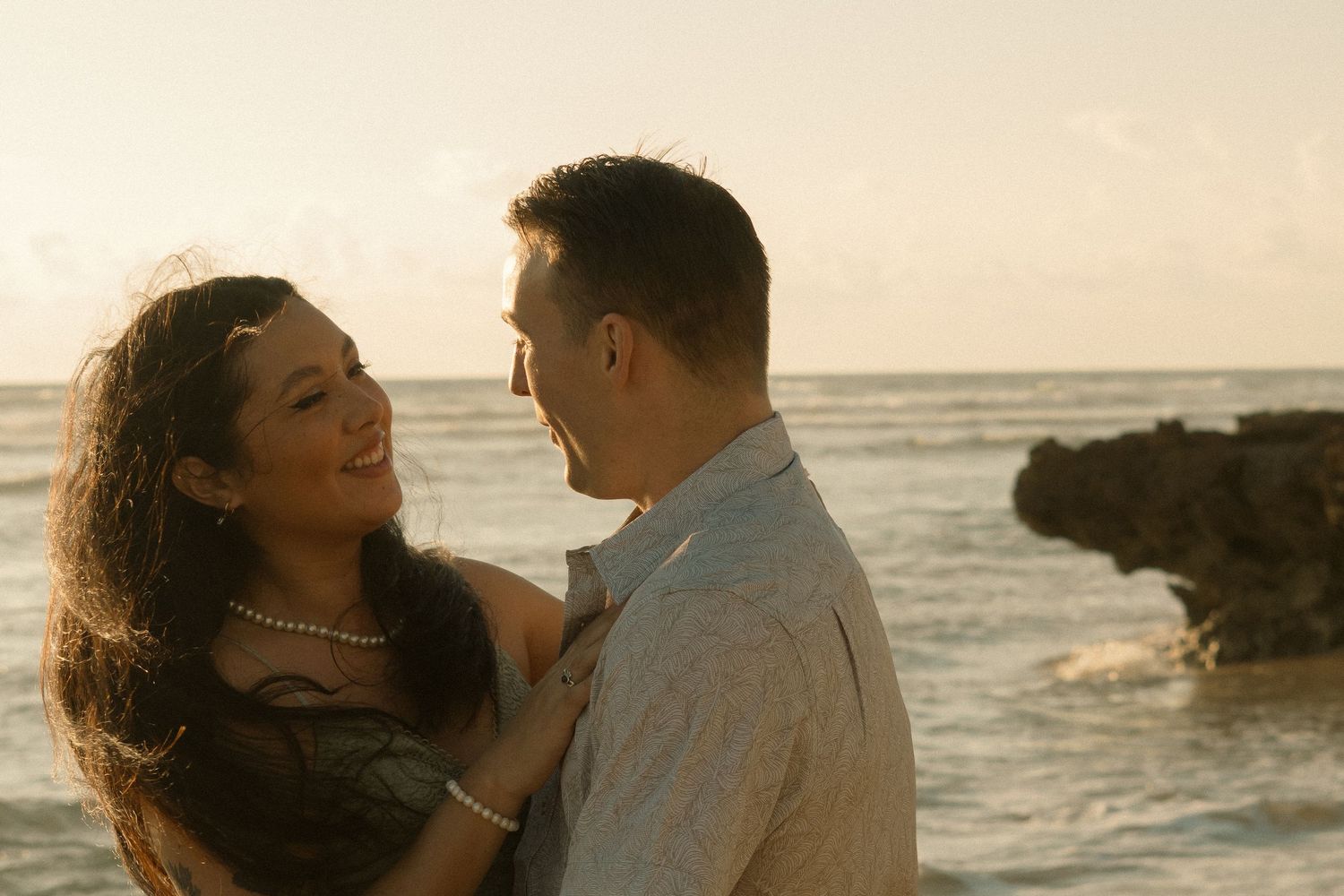 Couple sharing a romantic moment on a tropical beach at sunset with ocean waves and rocky shore in background.
