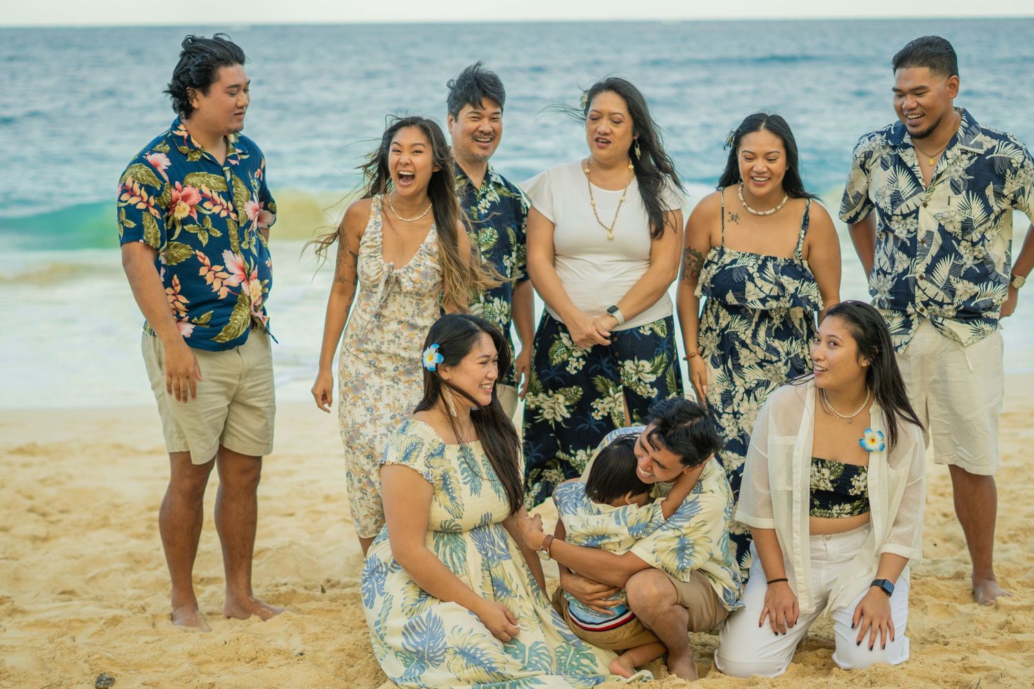 A joyful family group photo on a sandy beach, wearing matching Hawaiian floral outfits, laughing and posing together.