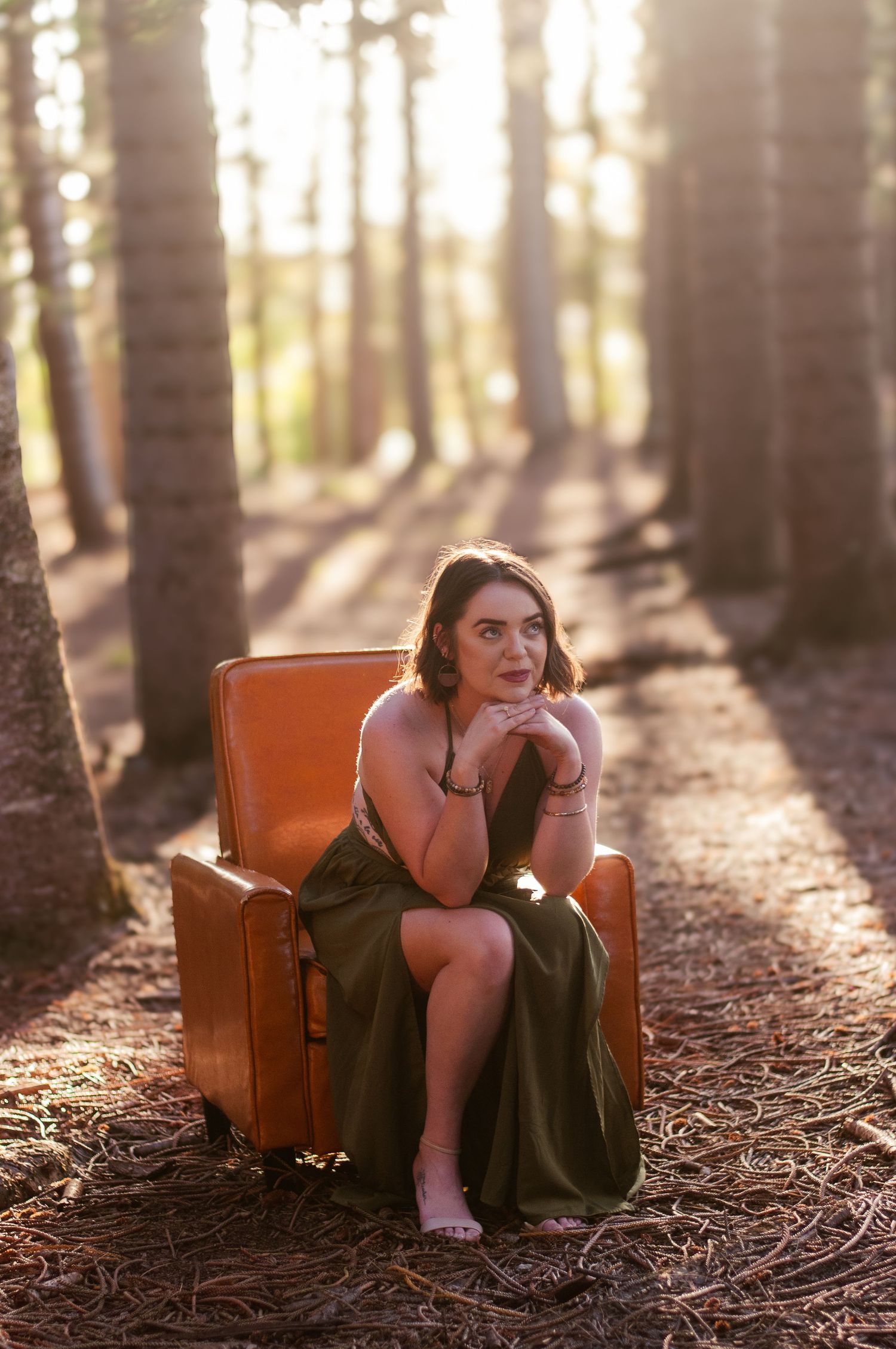 Young woman in olive green dress sitting in orange armchair in sunlit pine forest.