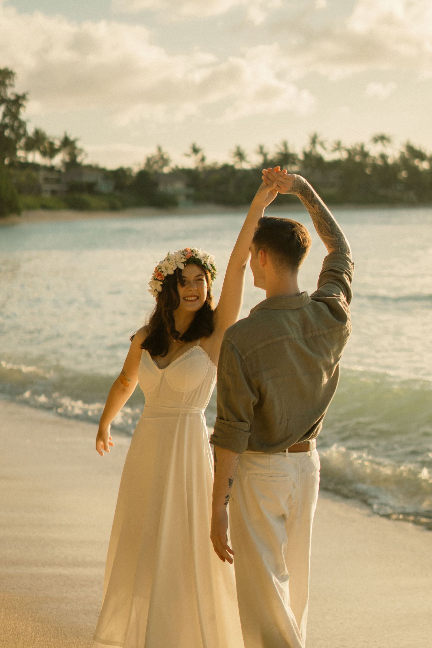 Bride in floral crown twirls in white dress as groom dances with her on Hawaii beach.
