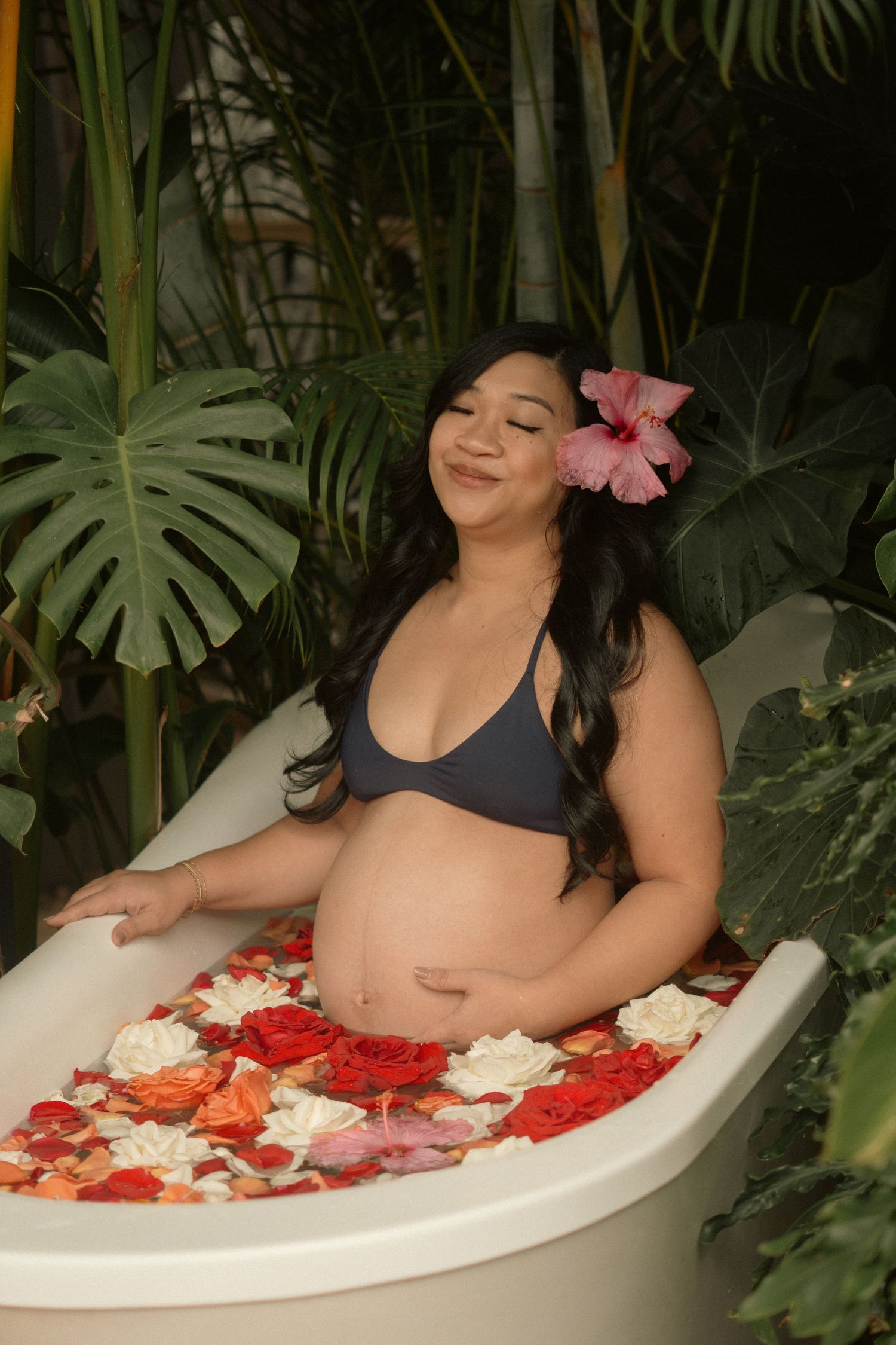 Pregnant woman relaxing in a floral bath surrounded by tropical plants, wearing a gray bikini top with a pink flower in her hair.