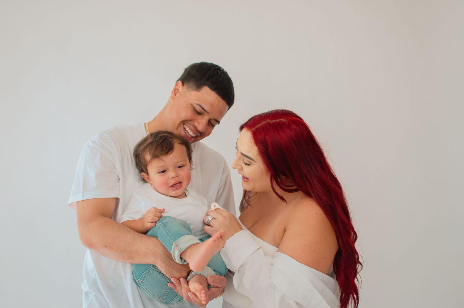 A smiling couple in white outfits lovingly gazes at their baby boy against a white background.