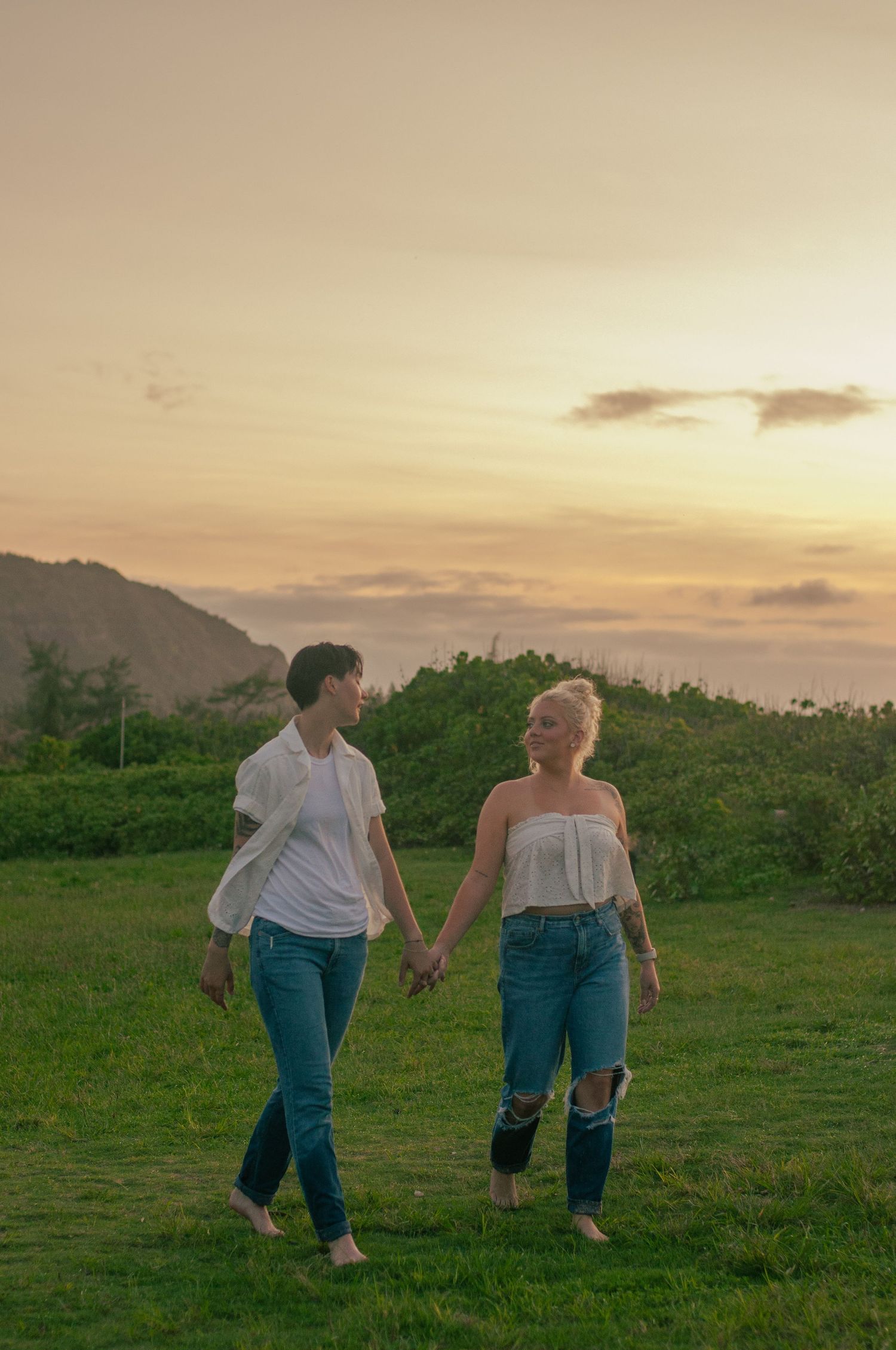 Couple holds hands walking barefoot on green hillside during golden sunset.